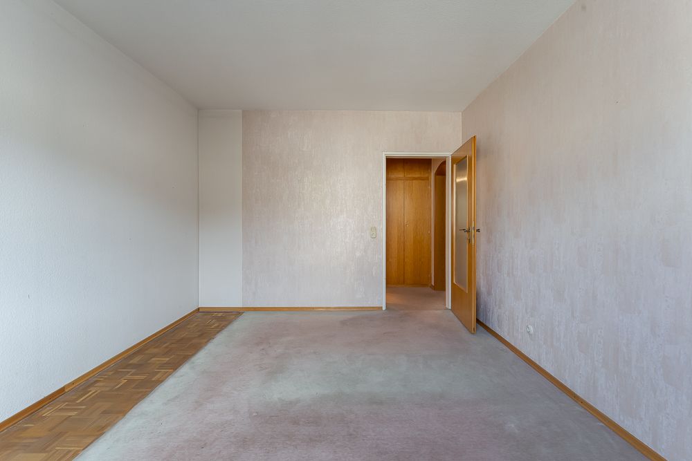 Leeres Zimmer mit hellem Teppichboden und offener Holztür zu einem Flur.