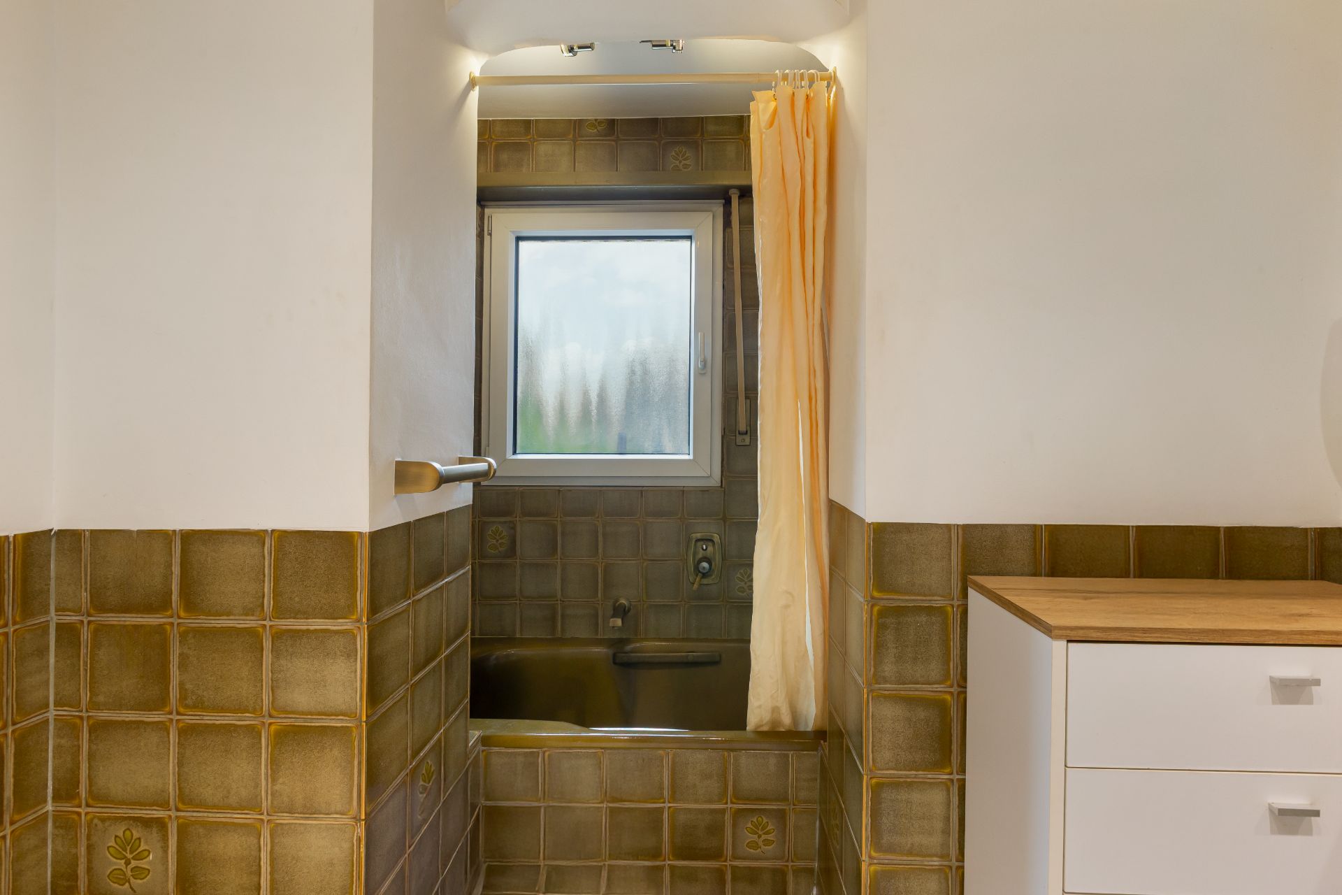 Bad mit braunen Fliesen, Badewanne, Fenster und beiger Duschvorhang.