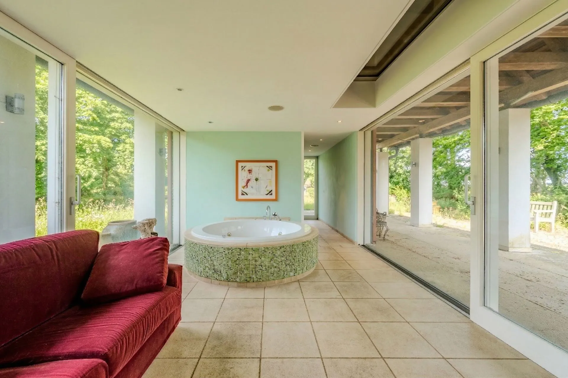 Heller Raum mit runder Badewanne, rotem Sofa und großen Fenstern mit Blick ins Grüne.