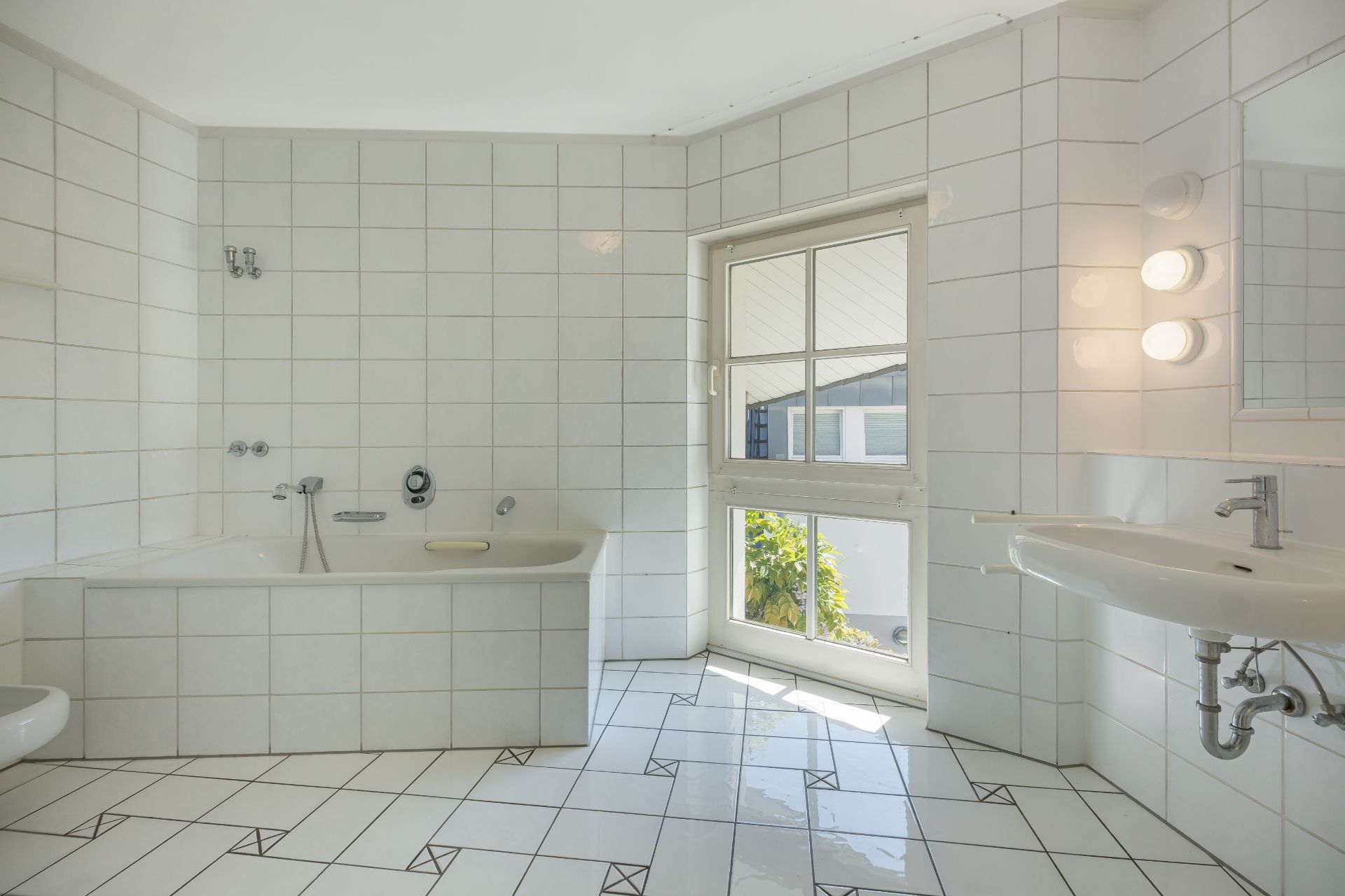 Helles Badezimmer mit Badewanne, Waschbecken und großem Fenster, weiße Fliesen an Wand und Boden.