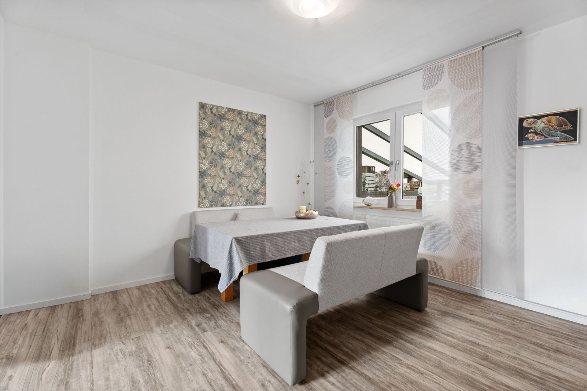 Essbereich mit grauer Eckbank, Tisch und Fenster, hellem Boden und Wanddekoration.