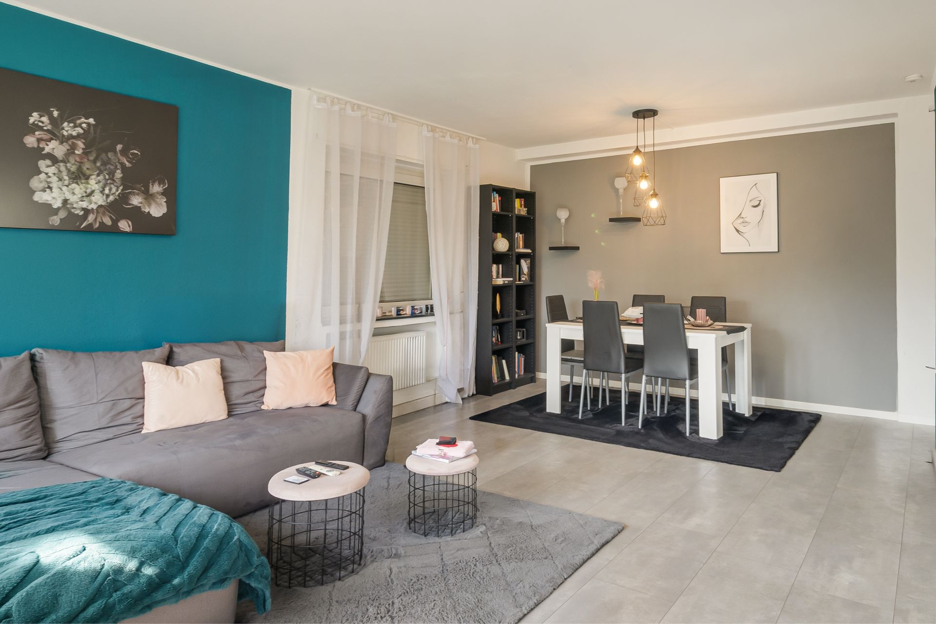 Modernes Wohnzimmer mit grauem Sofa, Esstisch, Bücherregal und Wandbildern.