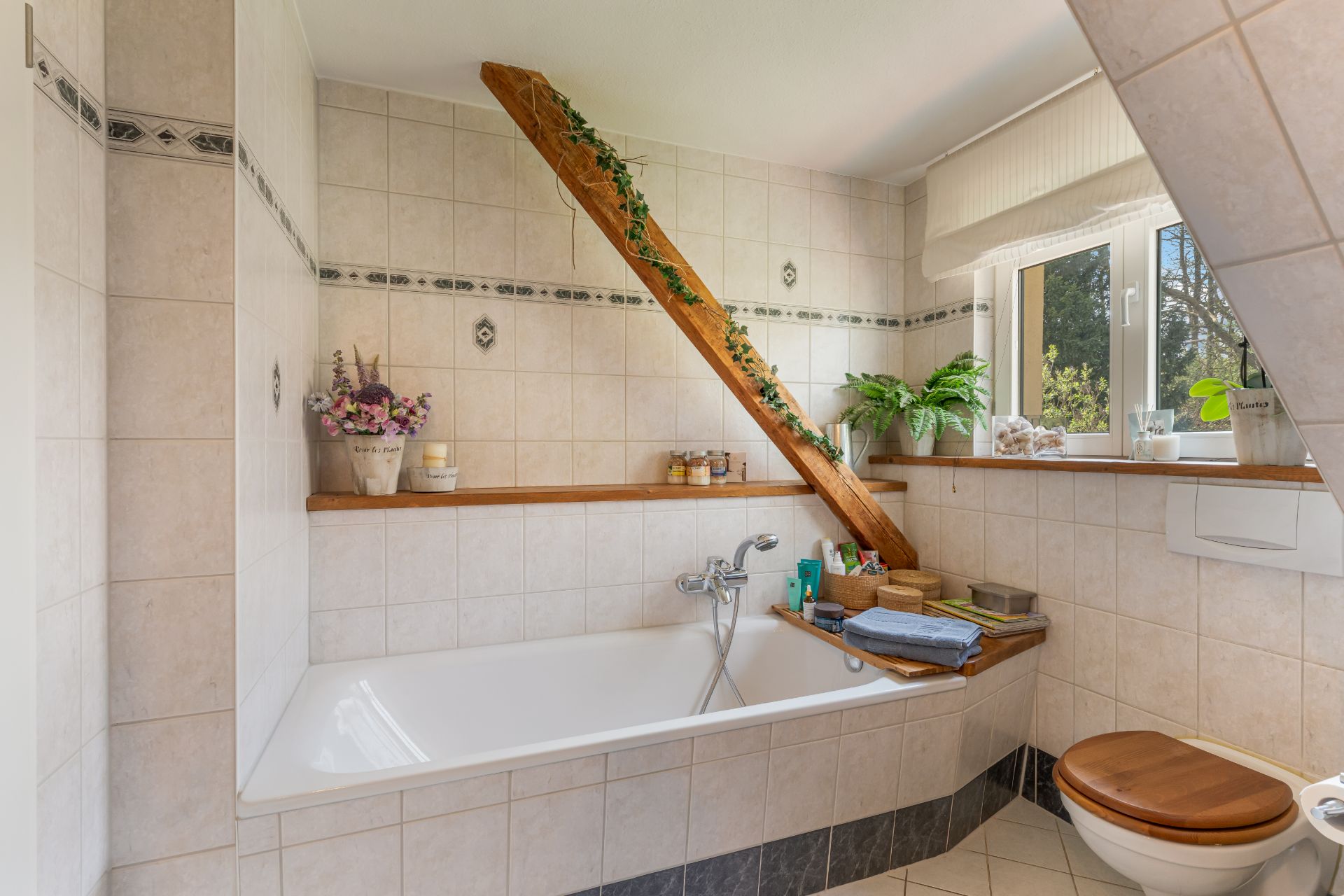 Helles Badezimmer mit Badewanne unter Dachschräge, Deko-Pflanzen und Fenster.