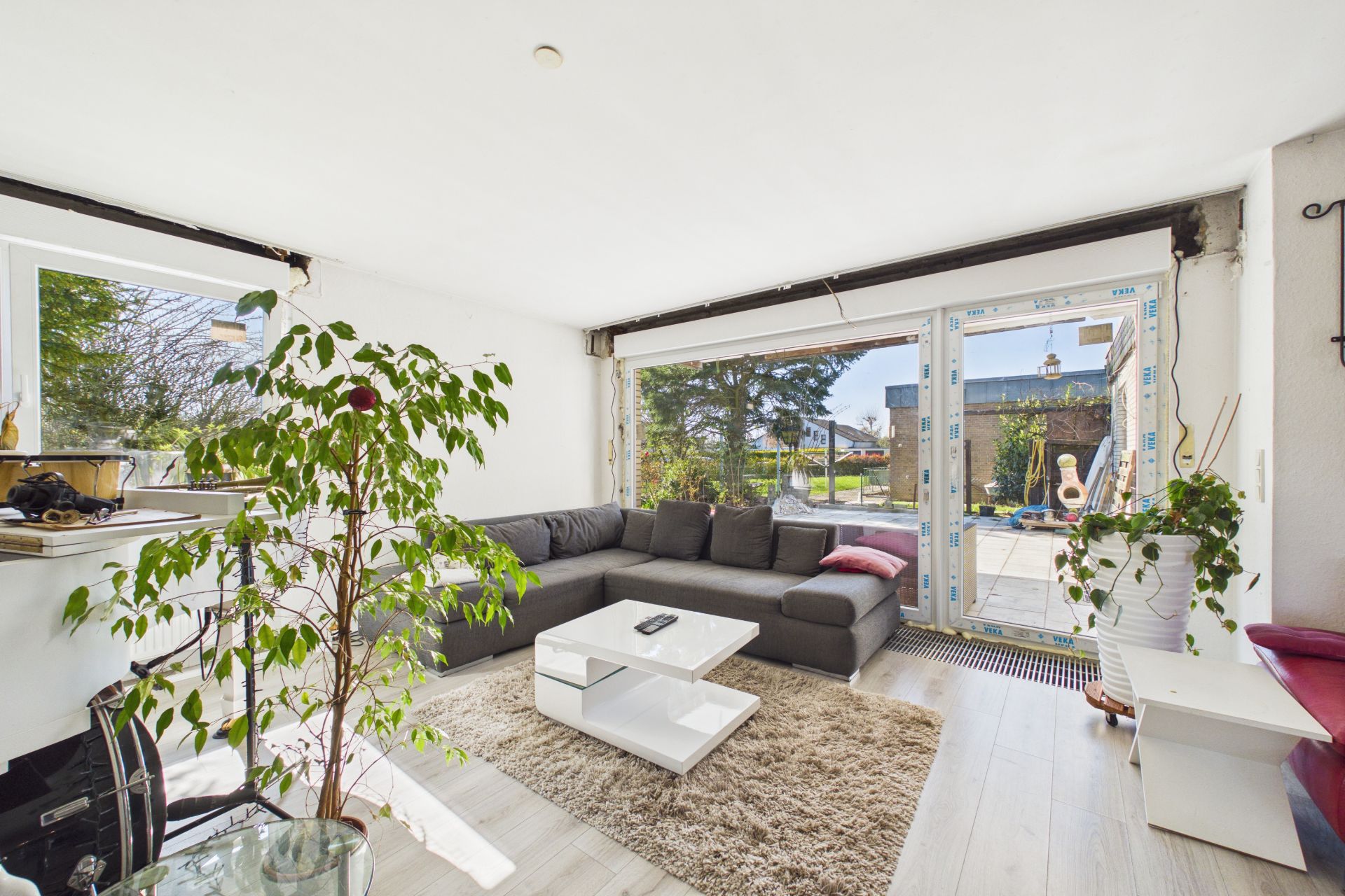 Helles Wohnzimmer mit großer Eckcouch, Couchtisch, Teppich und Zimmerpflanzen vor großen Fenstern.