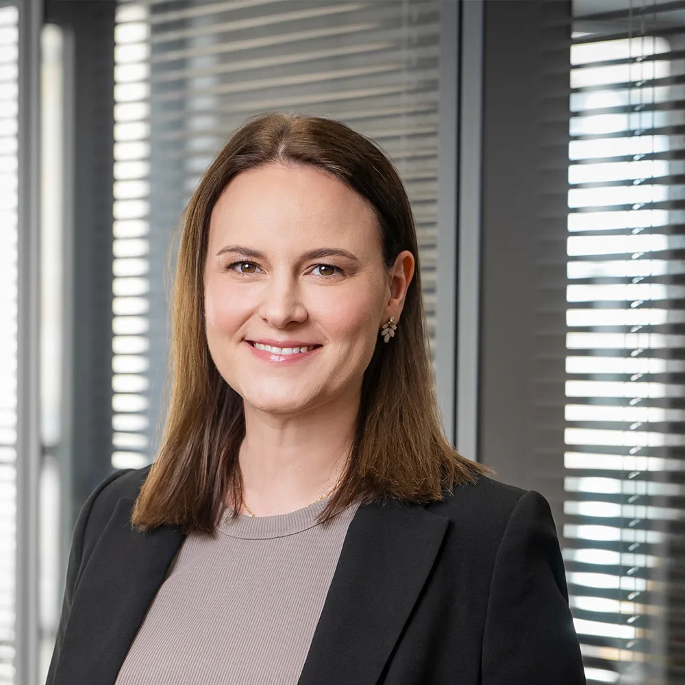 Frau mit braunen Haaren im Business-Outfit steht vor einem Fenster mit Jalousien.