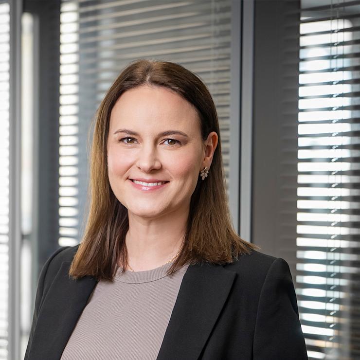 Frau mit braunen Haaren im Business-Outfit steht vor einem Fenster mit Jalousien.