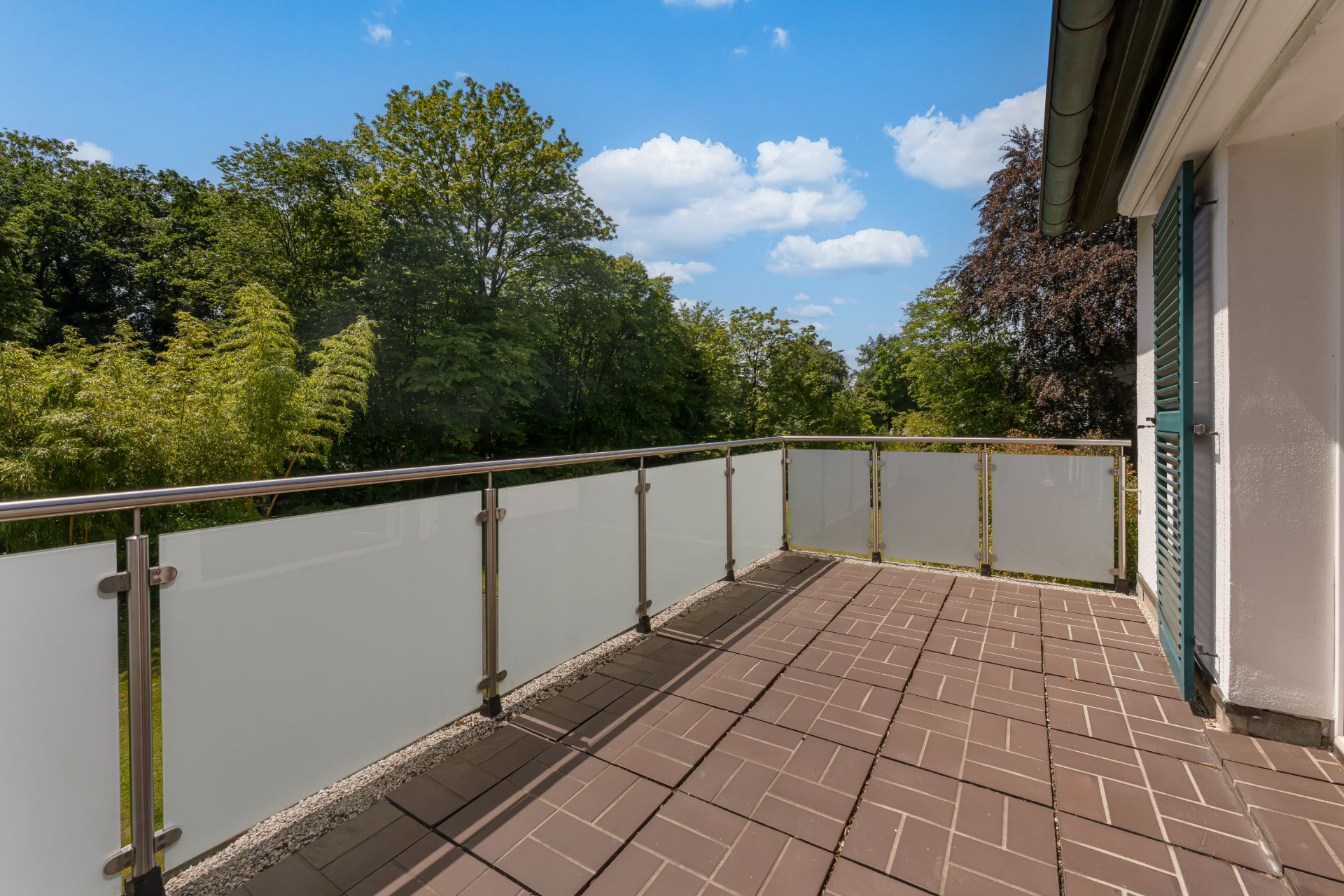 Großer Balkon mit braunen Fliesen und Glasgeländer, Blick auf grüne Bäume und blauen Himmel.