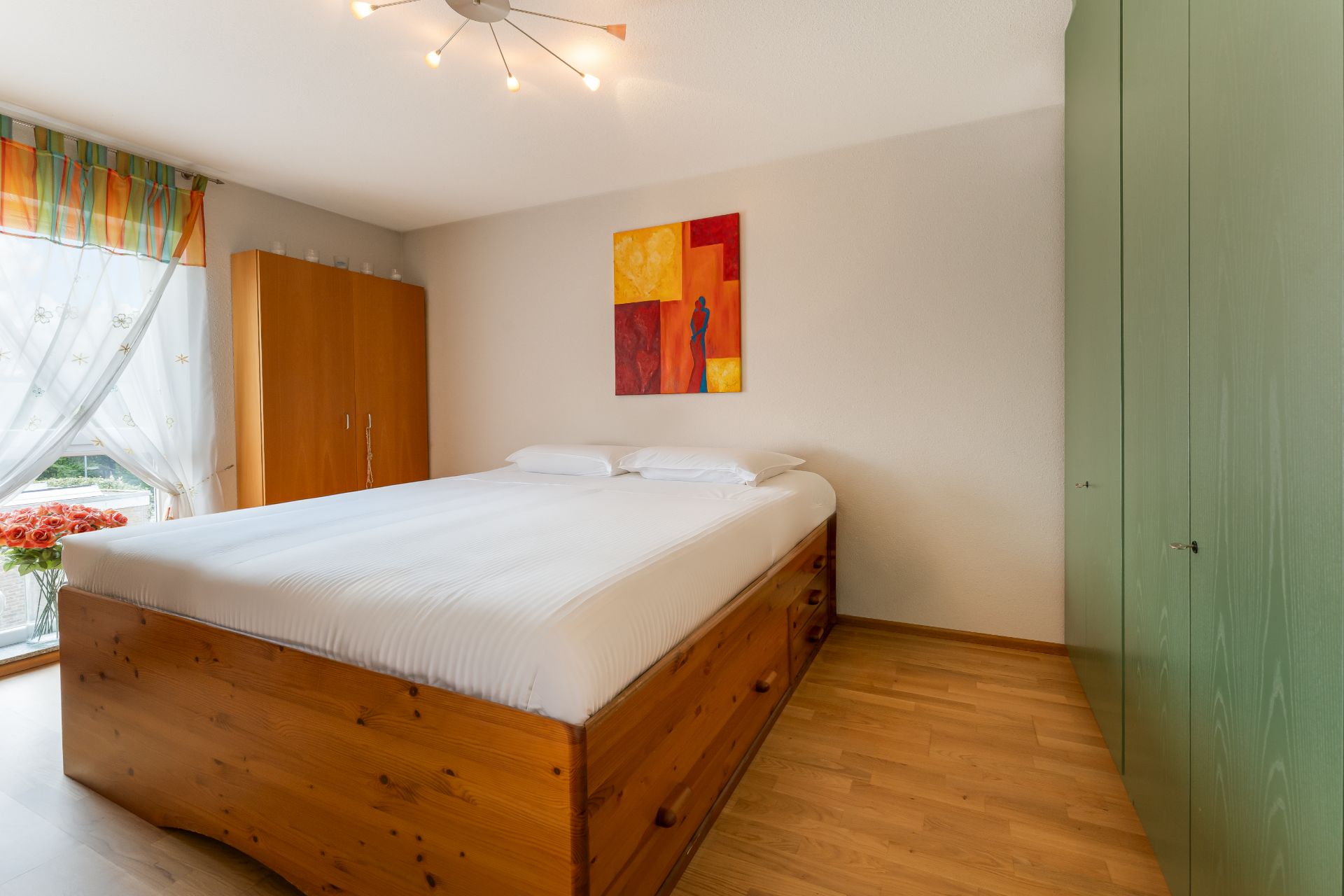 Helles Schlafzimmer mit Holzbett, grünem Schrank und buntem Bild an der Wand.