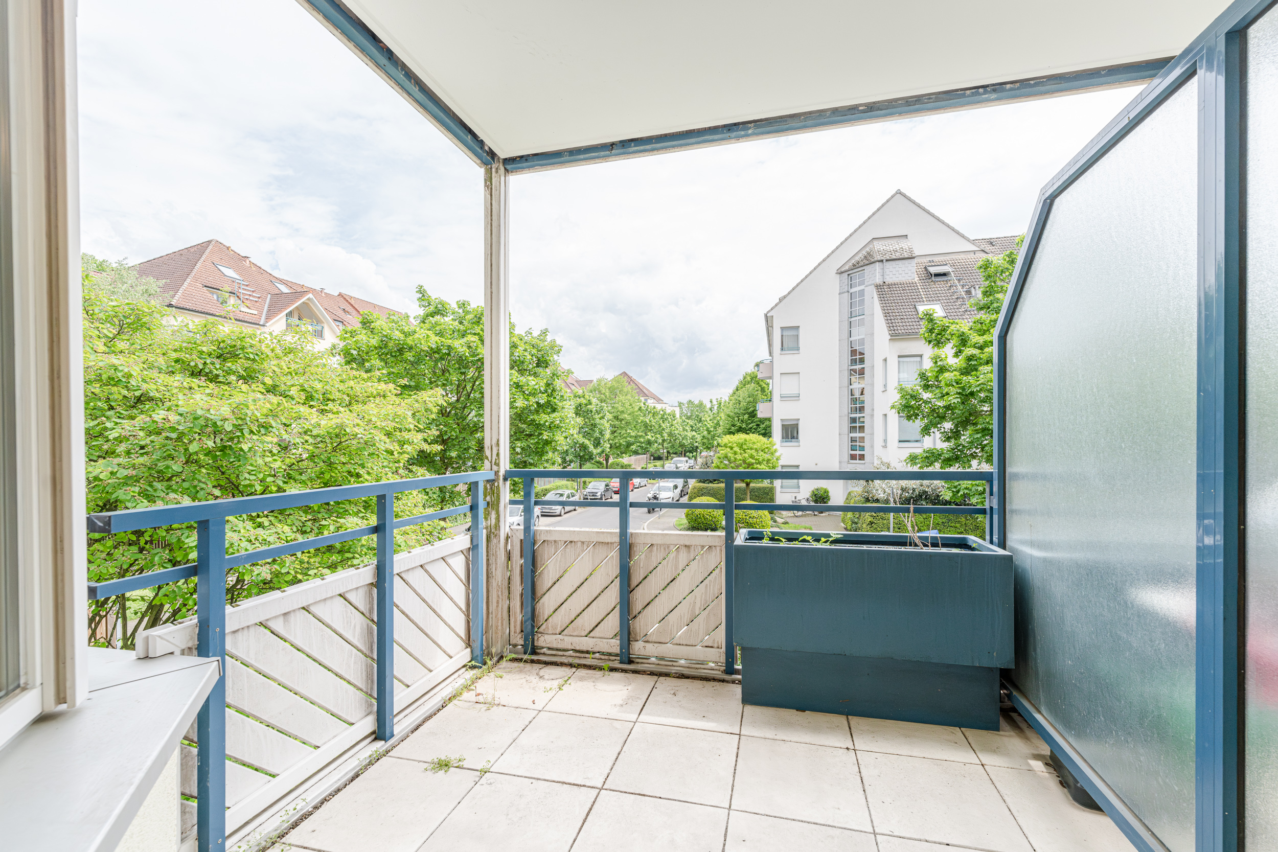 Überdachter Balkon mit blauem Geländer, Pflanzkasten und Blick auf grüne Bäume und Wohngebäude.