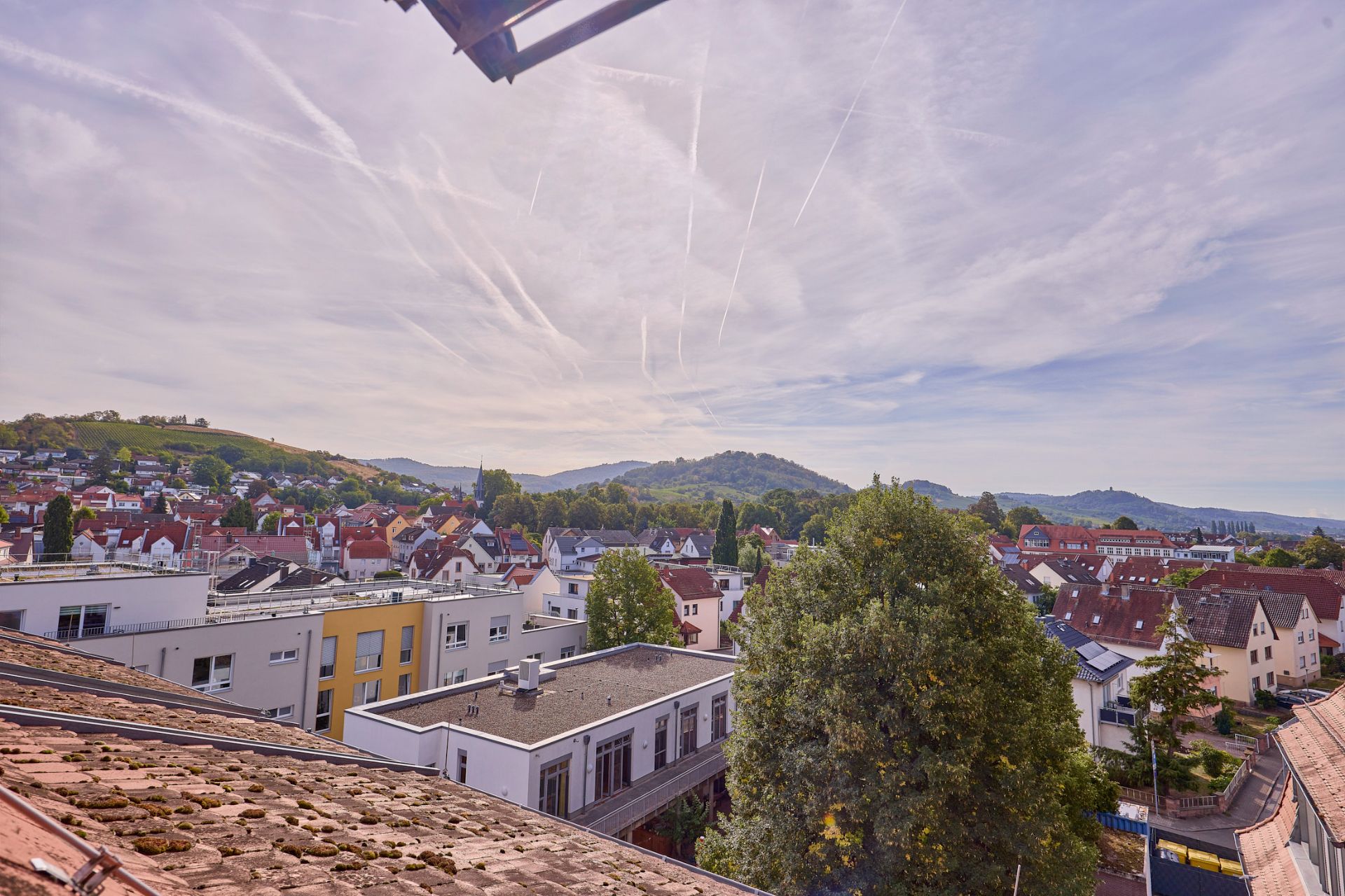 Blick über Wohnhäuser, Bäume und Hügel im Hintergrund unter leicht bewölktem Himmel.