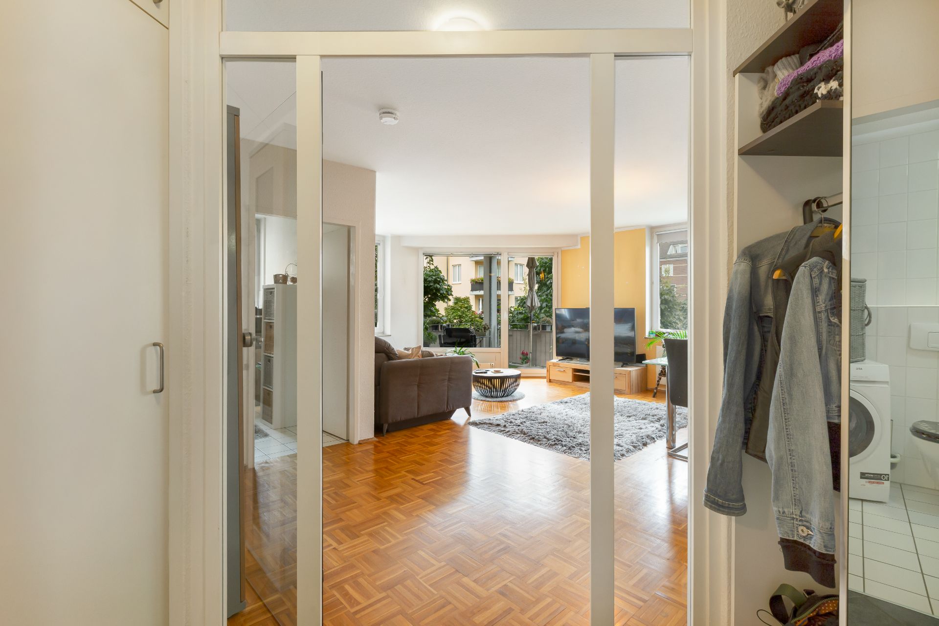 Blick durch Glastüren in ein helles Wohnzimmer mit Sofa, Fernseher und Balkon.