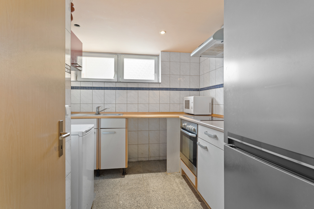 Kleine Küche mit Spüle, Herd, Mikrowelle und Kühlschrank, helle Fliesen und zwei Fenster.
