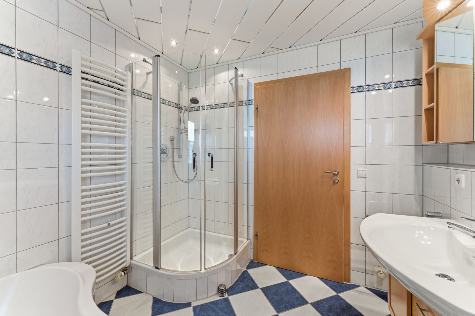 Modernes Bad mit Dusche, Waschbecken, Handtuchheizkörper und Holztür.