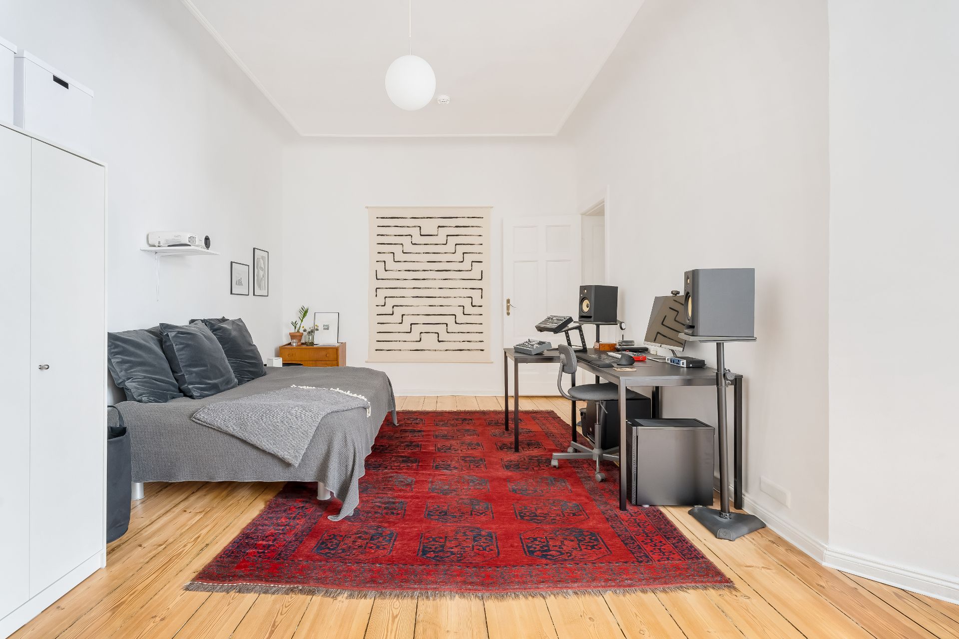 Helles Zimmer mit Bett, Schreibtisch mit Technik, rotem Teppich und Holzboden.