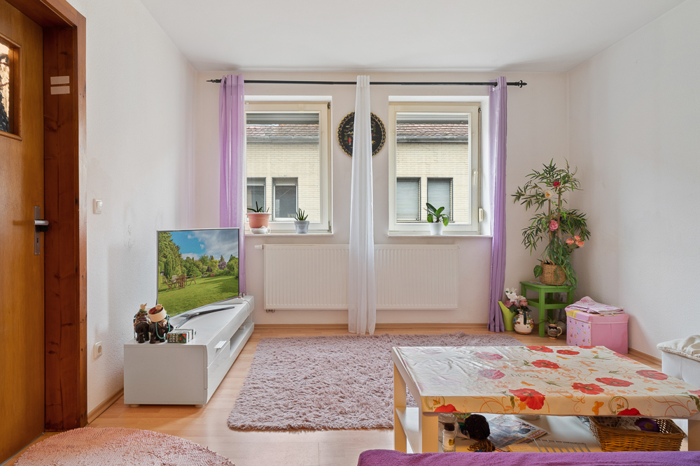 Wohnzimmer mit zwei Fenstern, lila Vorhängen, Fernseher, Teppich und Pflanzen.