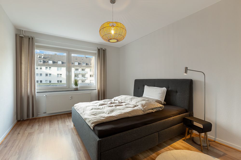 Helles Schlafzimmer mit großem Bett, Nachttisch, Stehlampe und Fenster mit Blick auf andere Gebäude.