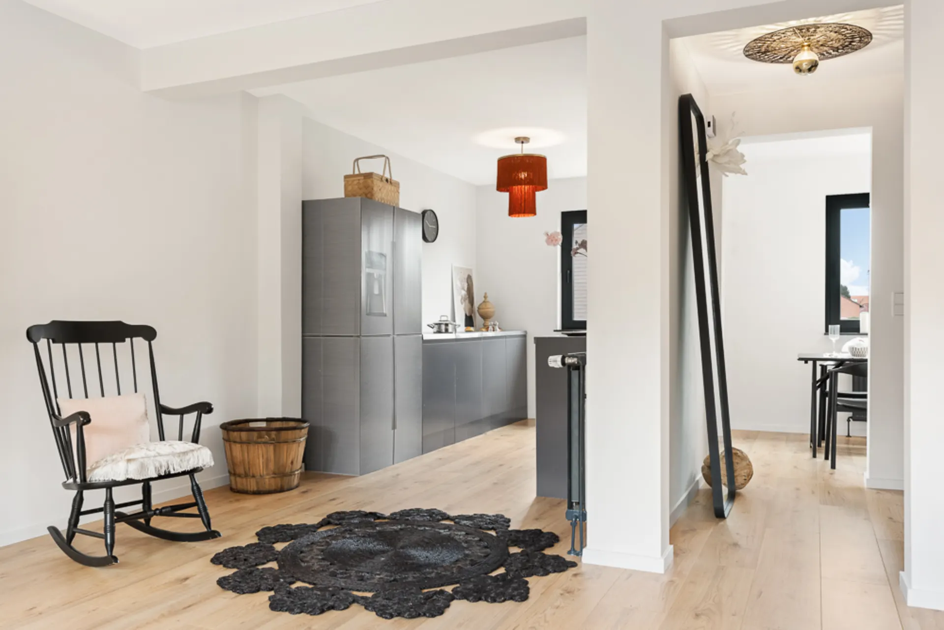 Helles Wohnzimmer mit Schaukelstuhl, Teppich, moderner Küche und Esstisch im Hintergrund.