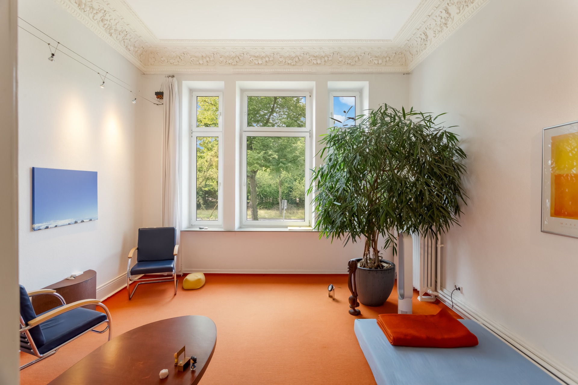 Helles Wohnzimmer mit großen Fenstern, zwei Stühlen, Pflanze, Tisch und Matte auf orangem Teppich.
