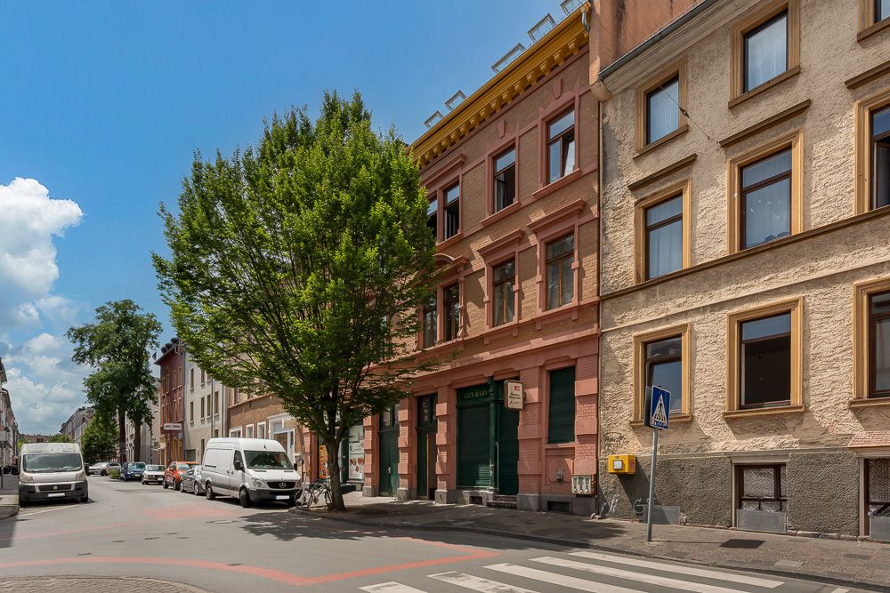Straßenszene mit Wohnhäusern, geparkten Autos und großem Baum am Straßenrand.