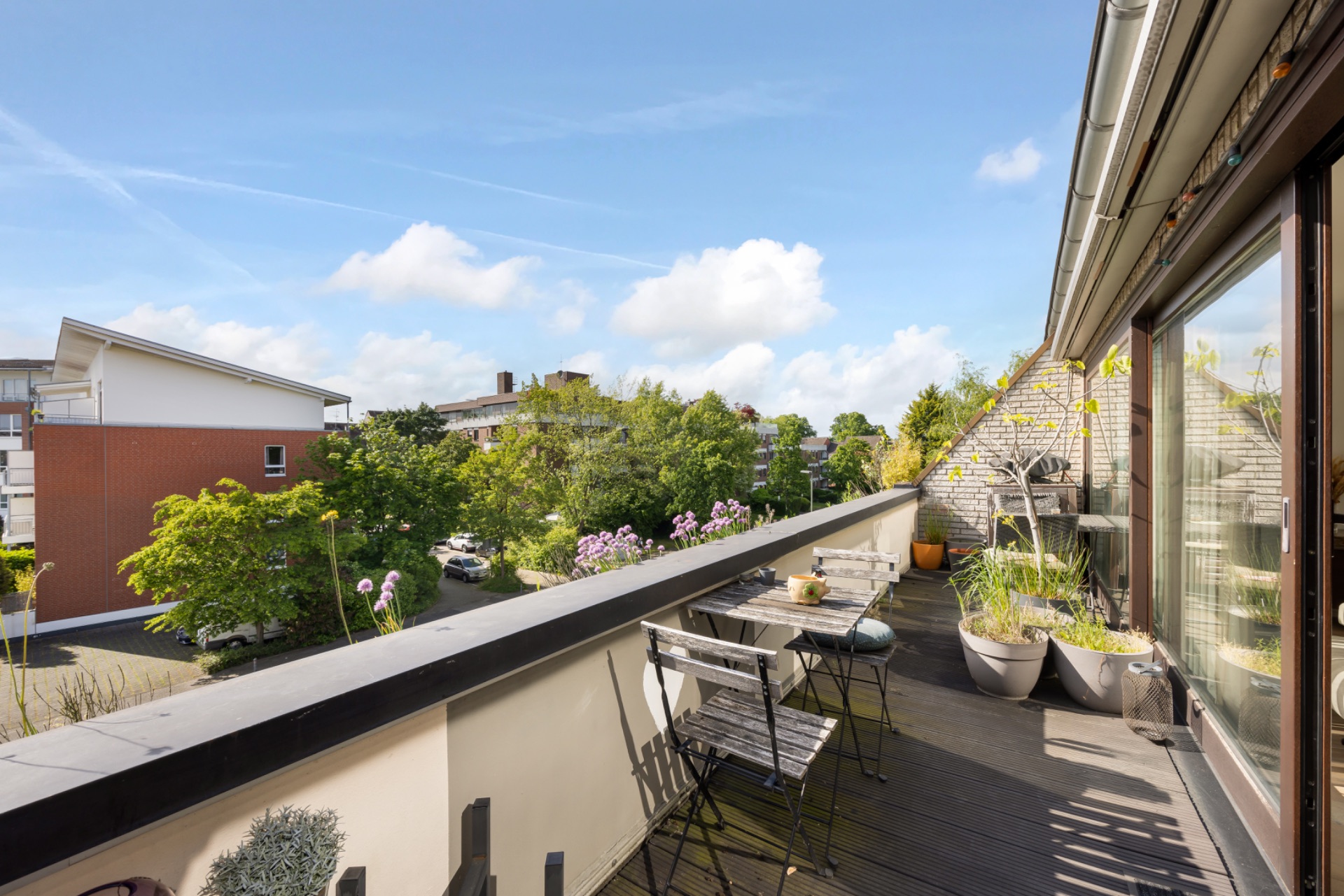Balkon mit Sitzgruppe und Pflanzen, Blick auf Nachbarhäuser und Bäume unter blauem Himmel.