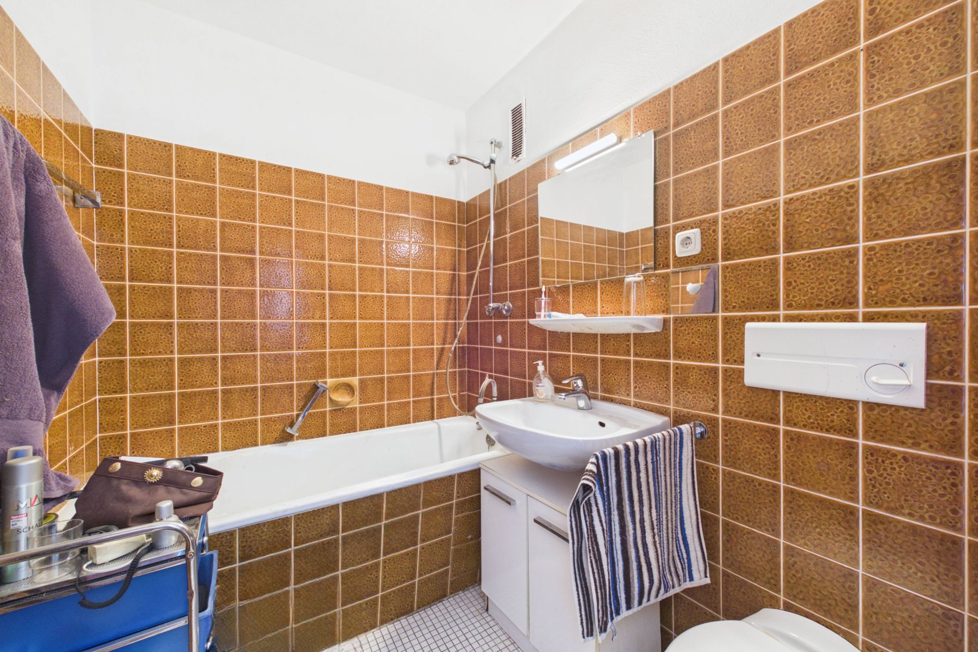 Kleines Bad mit braunen Fliesen, Badewanne, Waschbecken, Spiegel und Toilette.
