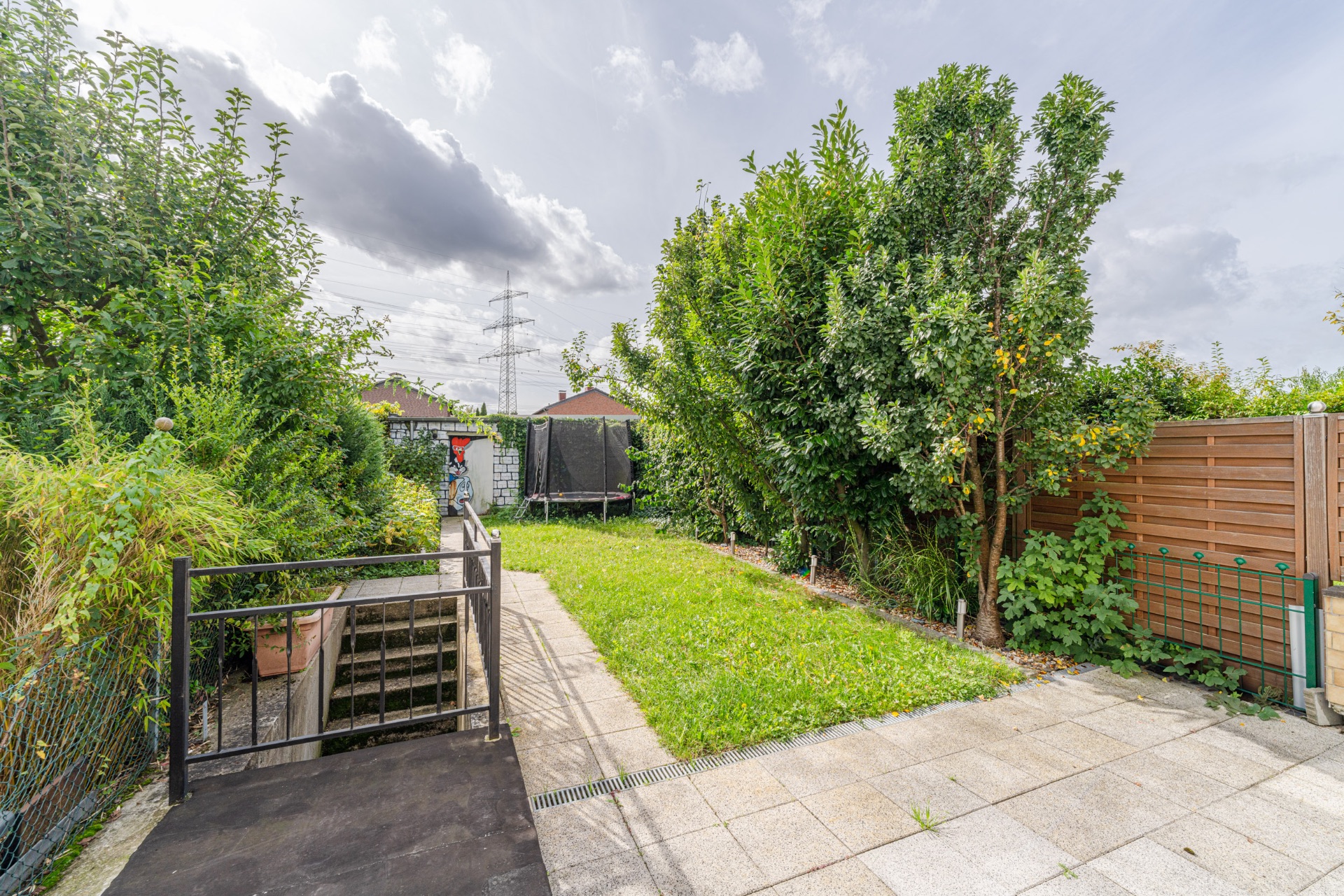 Garten mit Rasenfläche, Bäumen, Terrasse und Treppe, im Hintergrund ein Trampolin.