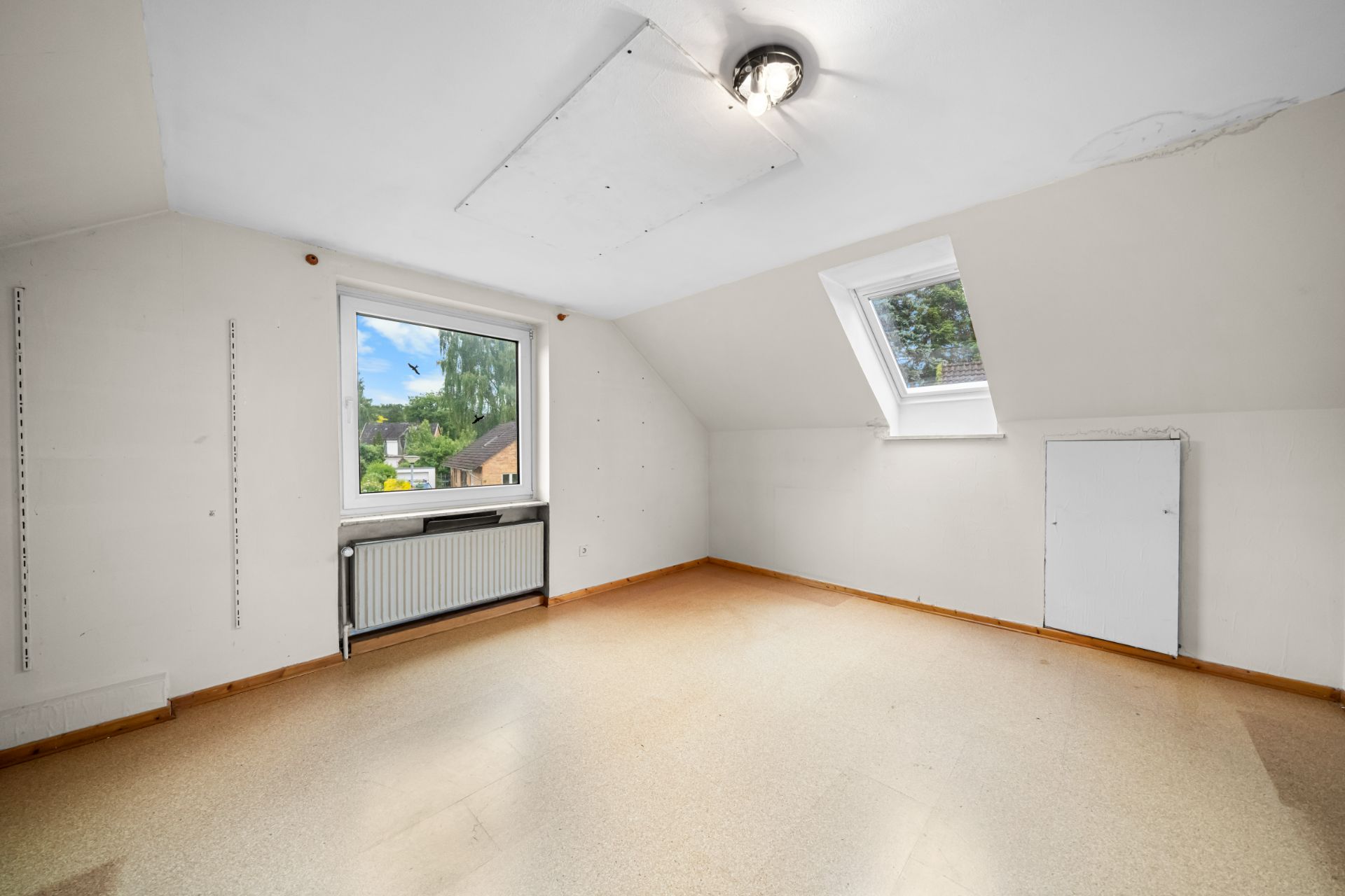 Helles, leeres Zimmer mit zwei Fenstern und Heizkörper unter einem Fenster.