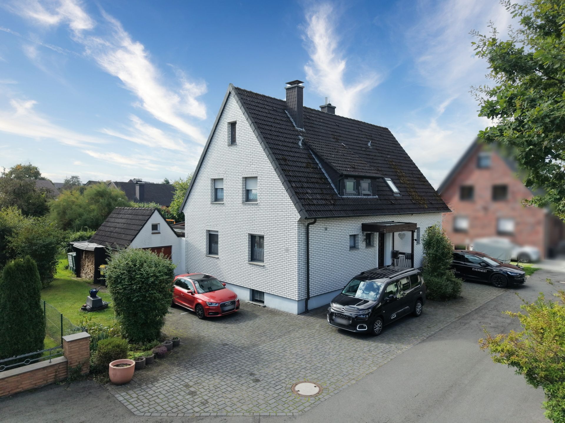 Weißes Einfamilienhaus mit dunklem Dach, drei Autos und kleinem Garten im Vordergrund.