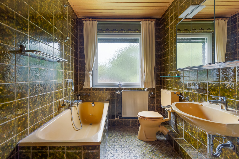Badezimmer mit gelber Badewanne, Toilette, Waschbecken und Fenster, grüne Fliesen an den Wänden.