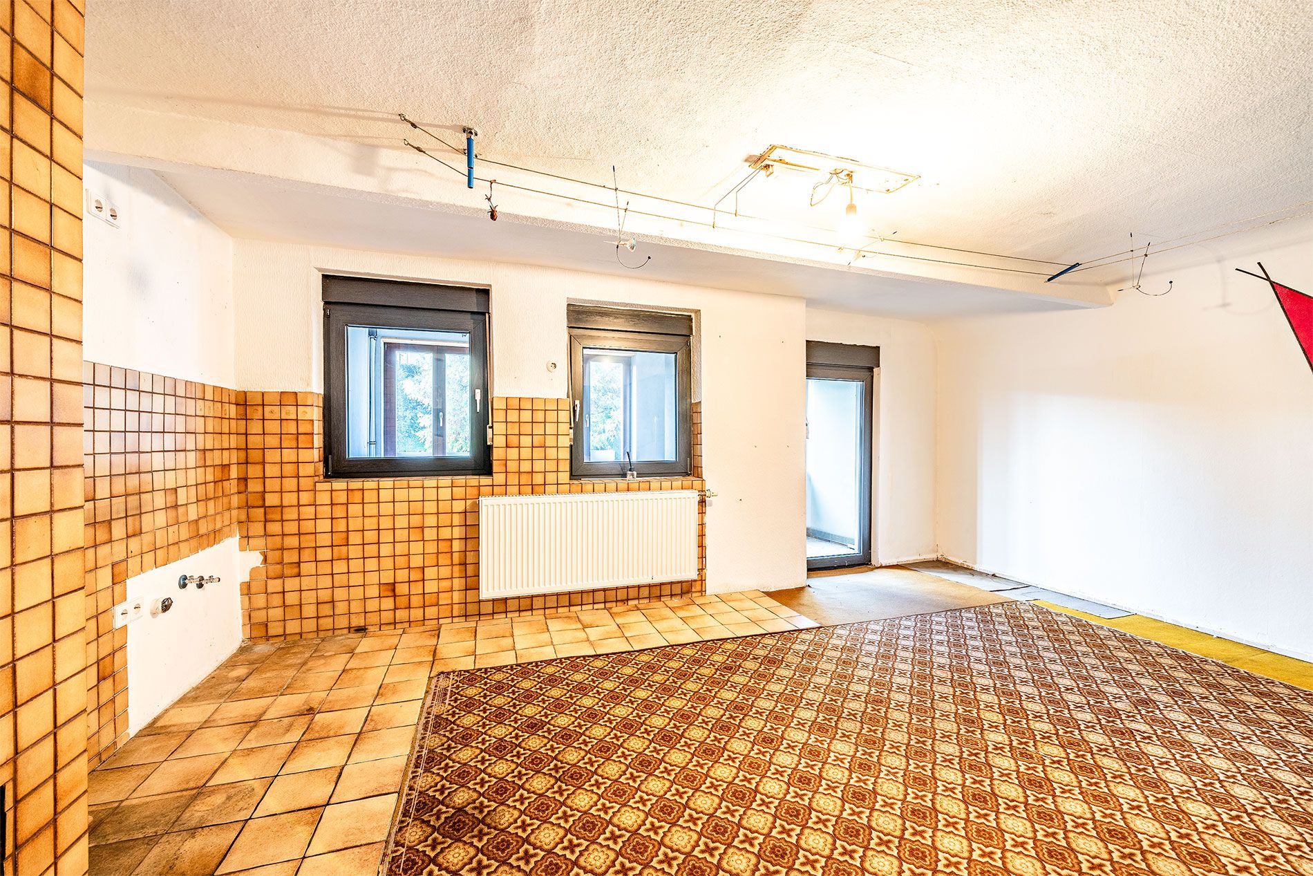Heller Raum mit zwei Fenstern, Fliesenboden, Teppichfläche und orangefarbenen Wandfliesen.