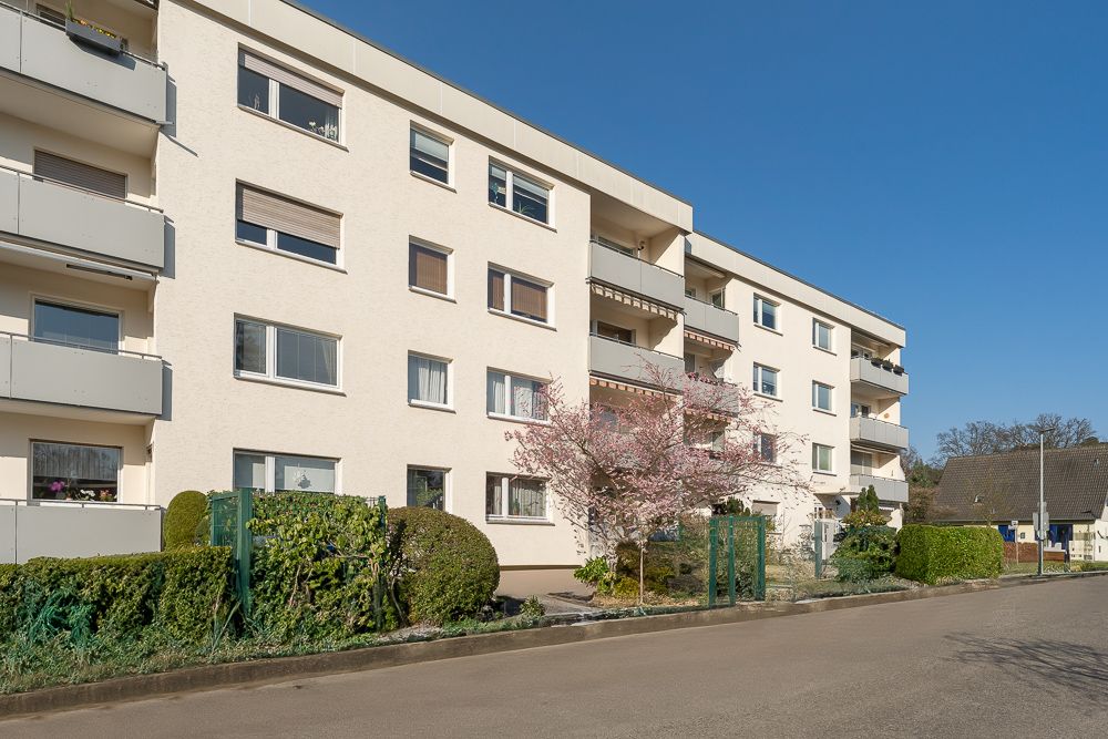 Mehrstöckiges Wohngebäude mit Balkonen und gepflegten Hecken an einer Straße.