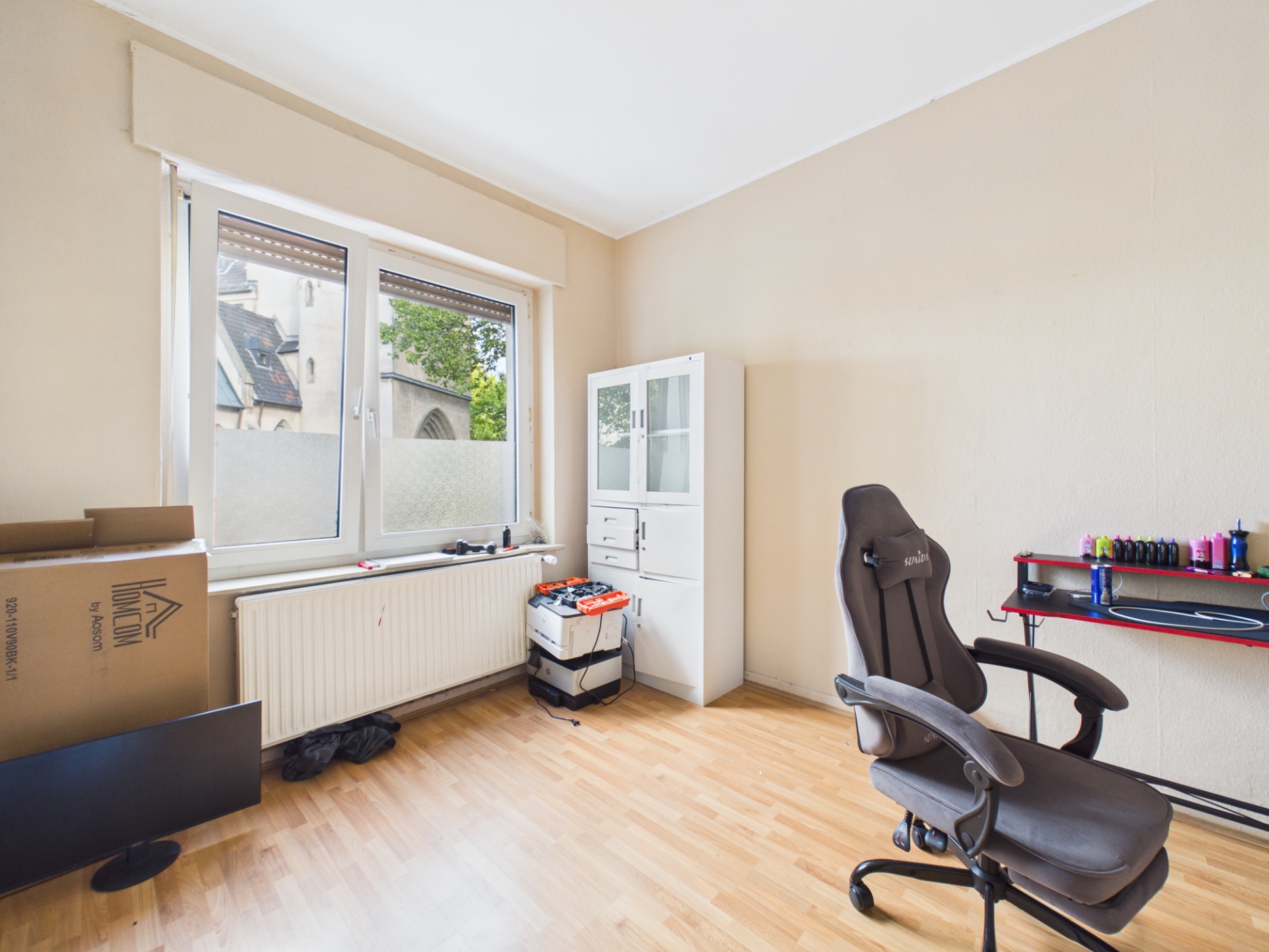 Helles Zimmer mit großem Fenster, Bürostuhl, Regal, Kartons und Holzboden.
