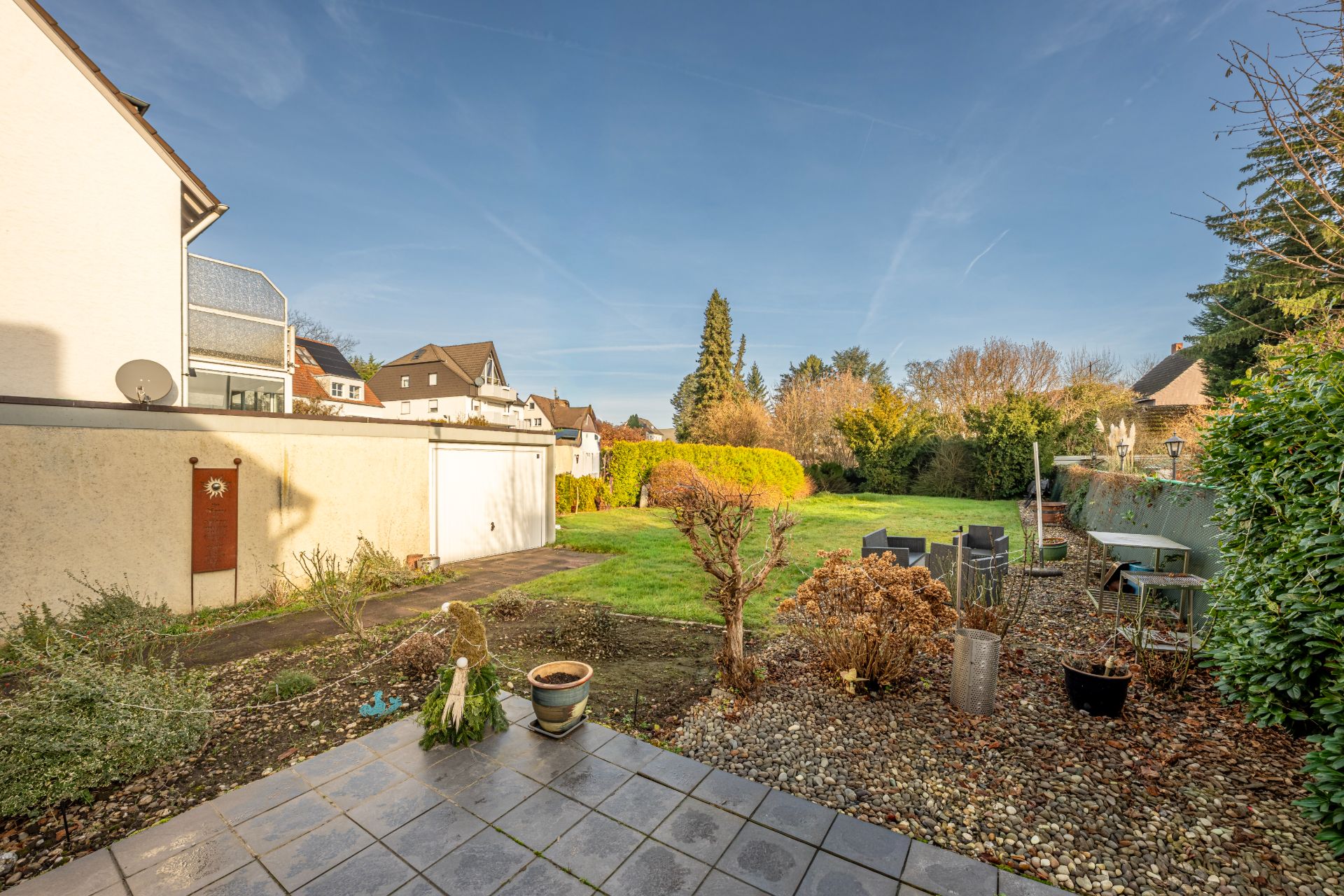 Garten mit Terrasse, Rasenfläche, Gartenmöbeln und Sträuchern an einem sonnigen Tag.