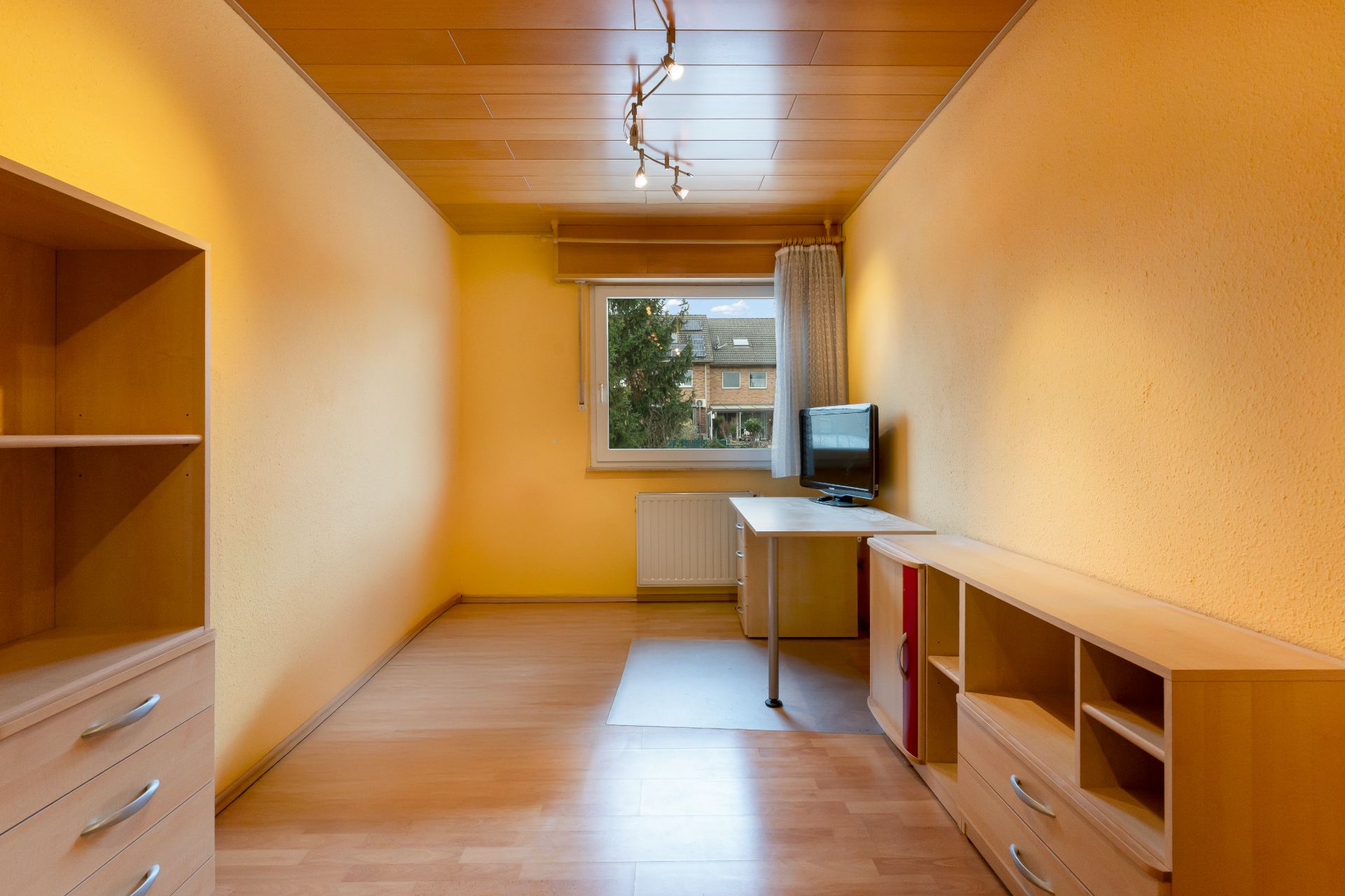 Schmaler Raum mit Holzregalen, Schreibtisch und Fenster, gelbe Wände und Holzdecke.