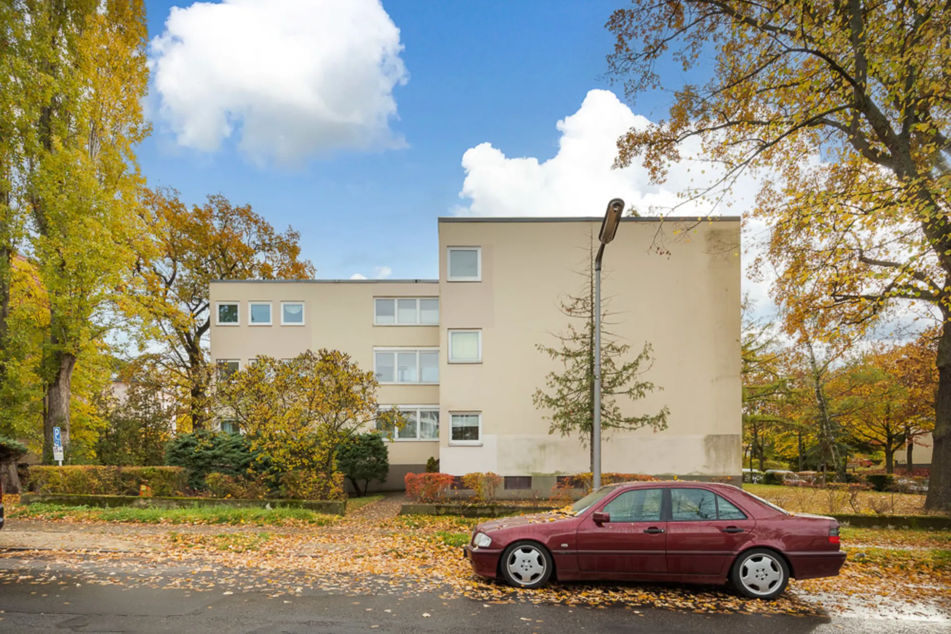 Wohnhaus mit mehreren Fenstern, rotes Auto parkt davor, Bäume mit Herbstlaub im Vordergrund.