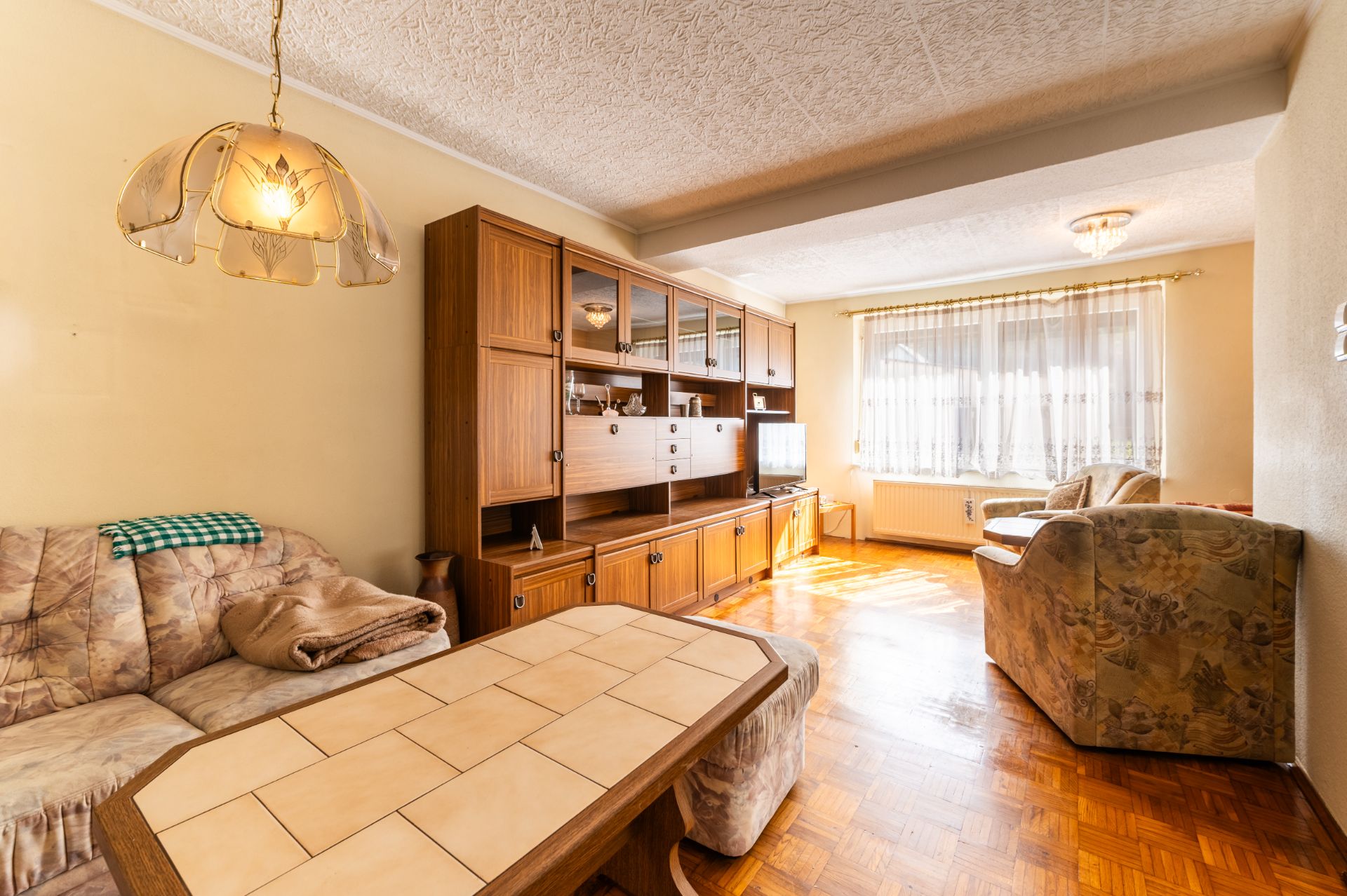 Helles Wohnzimmer mit Holzschrankwand, Sofas, Couchtisch und großem Fenster mit Gardinen.