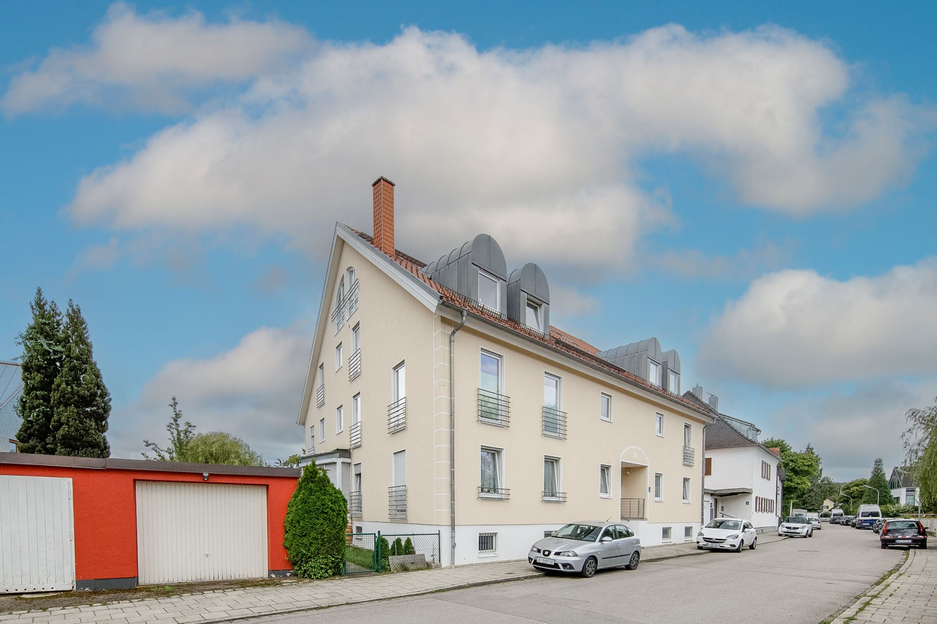 Mehrfamilienhaus mit roten Garagen und geparkten Autos an einer ruhigen Wohnstraße.
