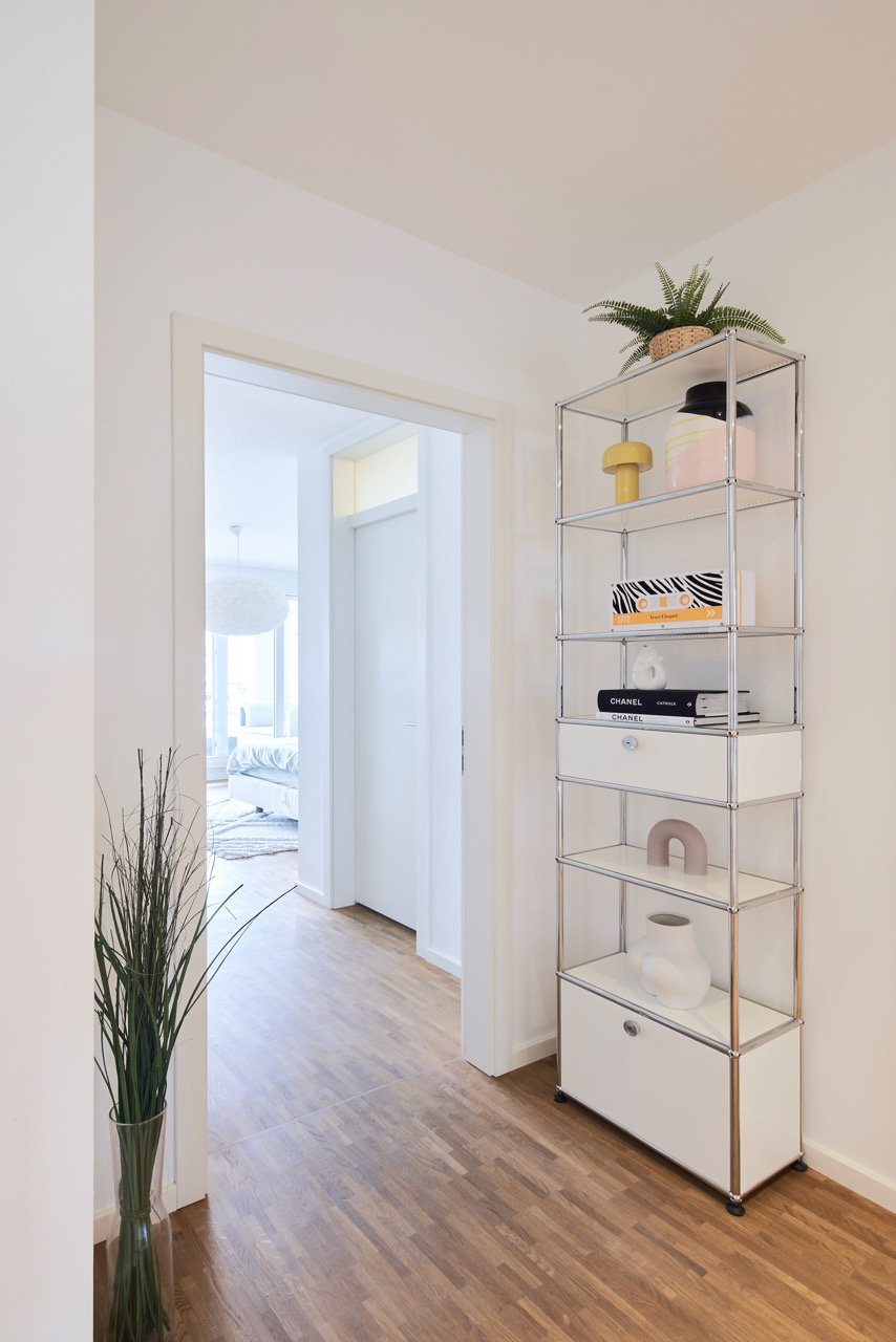 Heller Flur mit Holzboden, Metallregal und Blick ins Schlafzimmer.