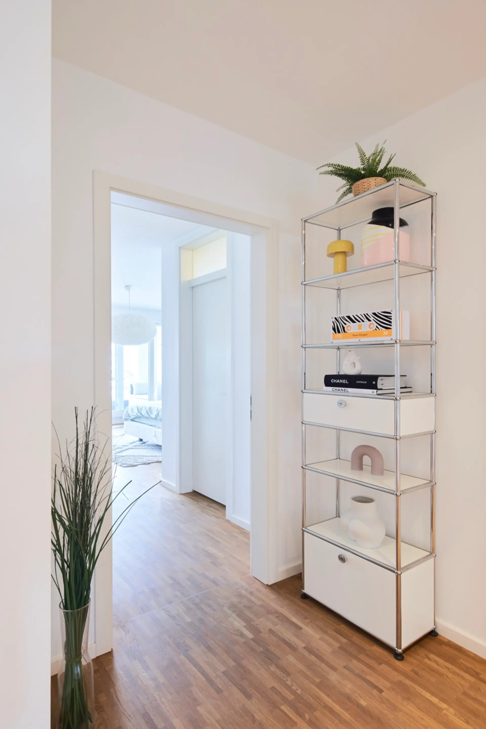 Heller Flur mit Holzboden, Metallregal und Blick ins Schlafzimmer.