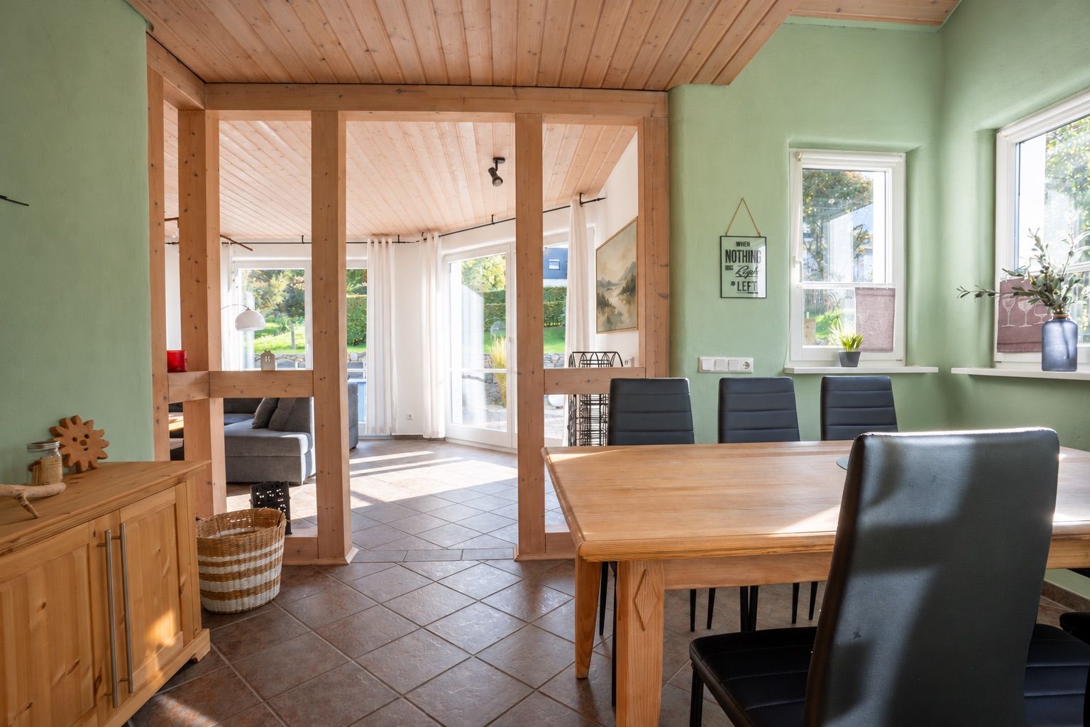 Esszimmer mit Holztisch, schwarzen Stühlen und Zugang zu einem sonnigen Wohnzimmer mit großen Fenstern.