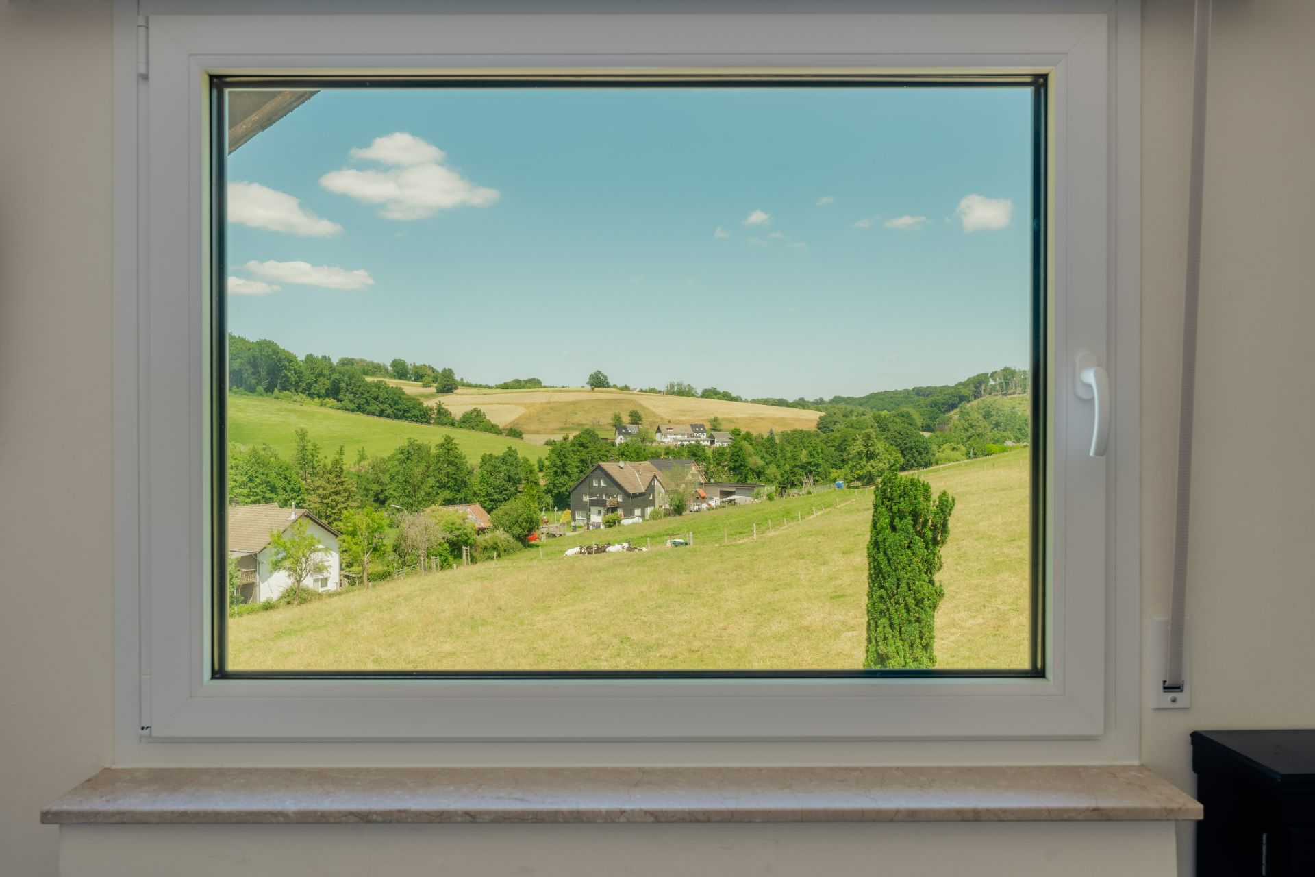 Blick durch ein Fenster auf grüne Hügel, Felder und ein paar Häuser im Dorf.