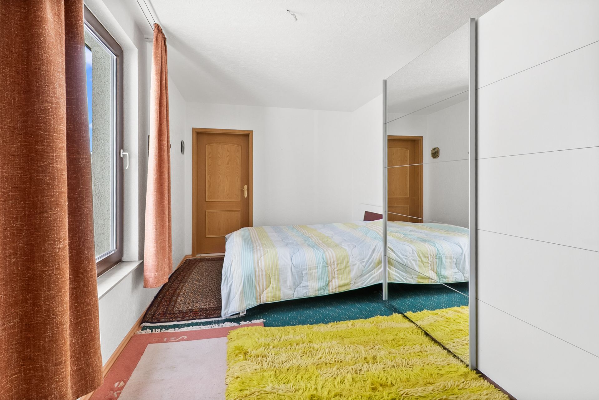 Schlafzimmer mit großem Fenster, Doppelbett, Spiegelschrank und bunten Teppichen.