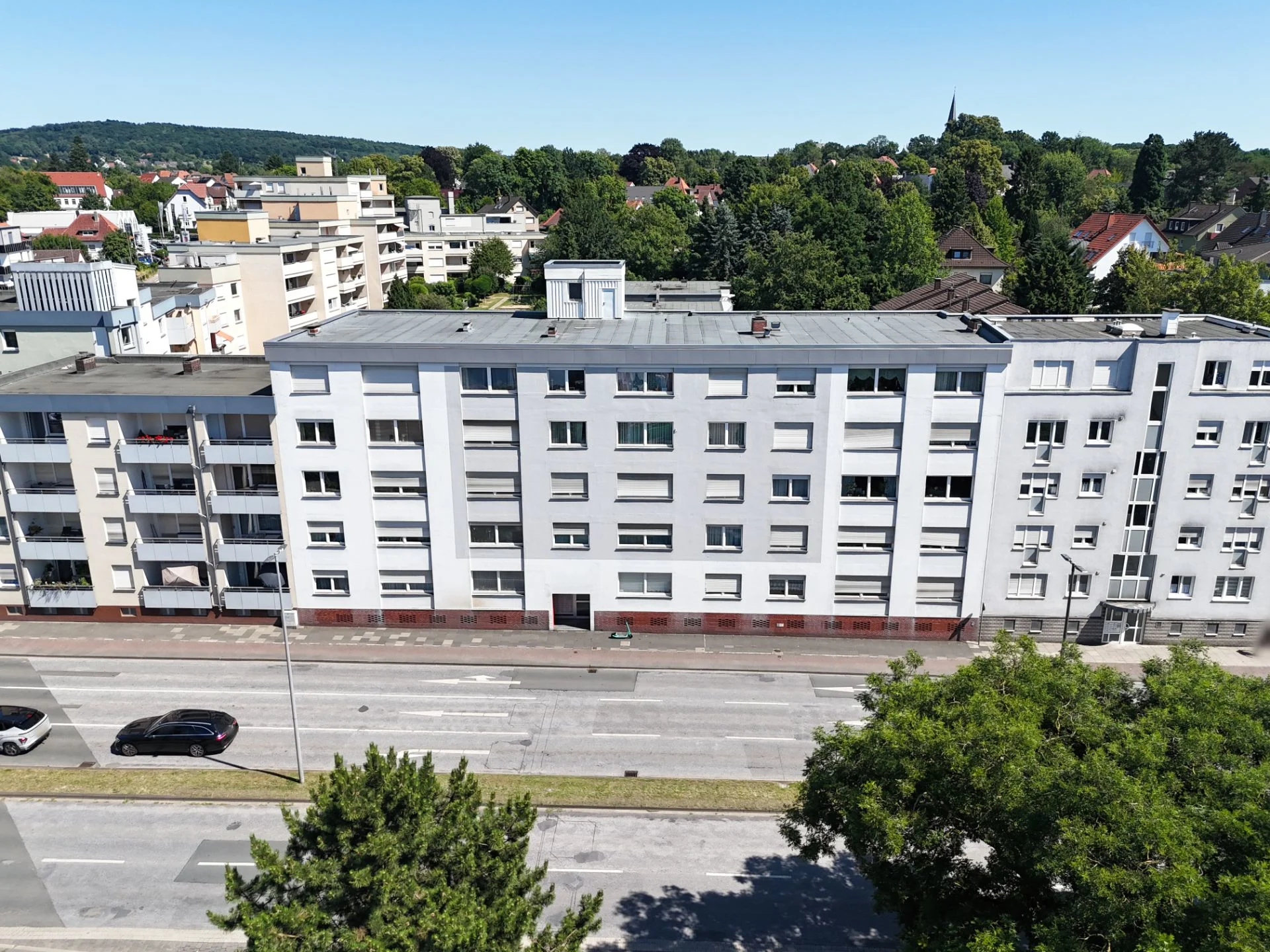Mehrstöckiges Wohnhaus mit Balkonen an einer Straße, umliegende Bäume und weitere Gebäude im Hintergrund.