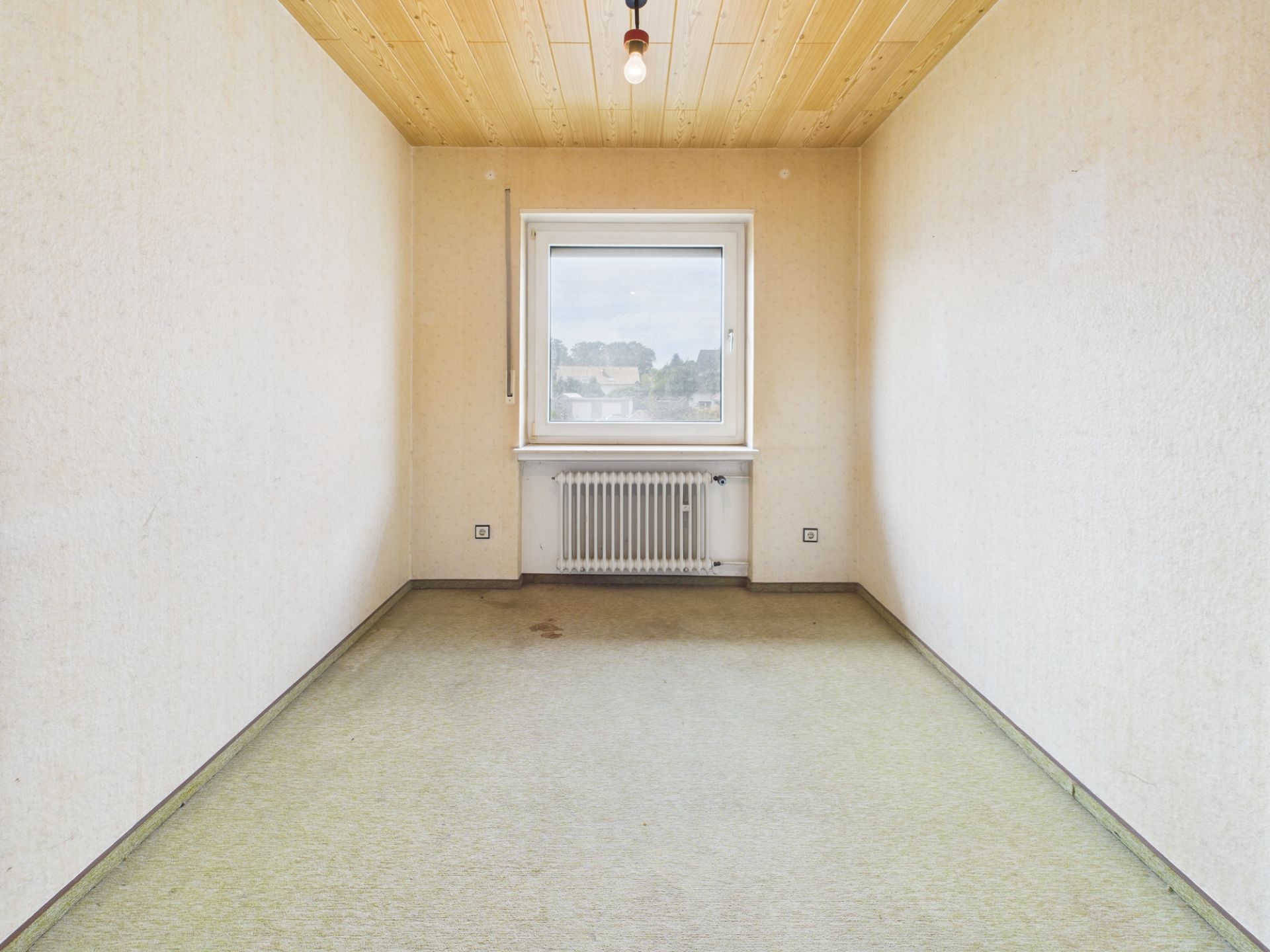 Leeres Zimmer mit Fenster, Heizkörper und Holzdecke, beige Wände und grünem Teppichboden.