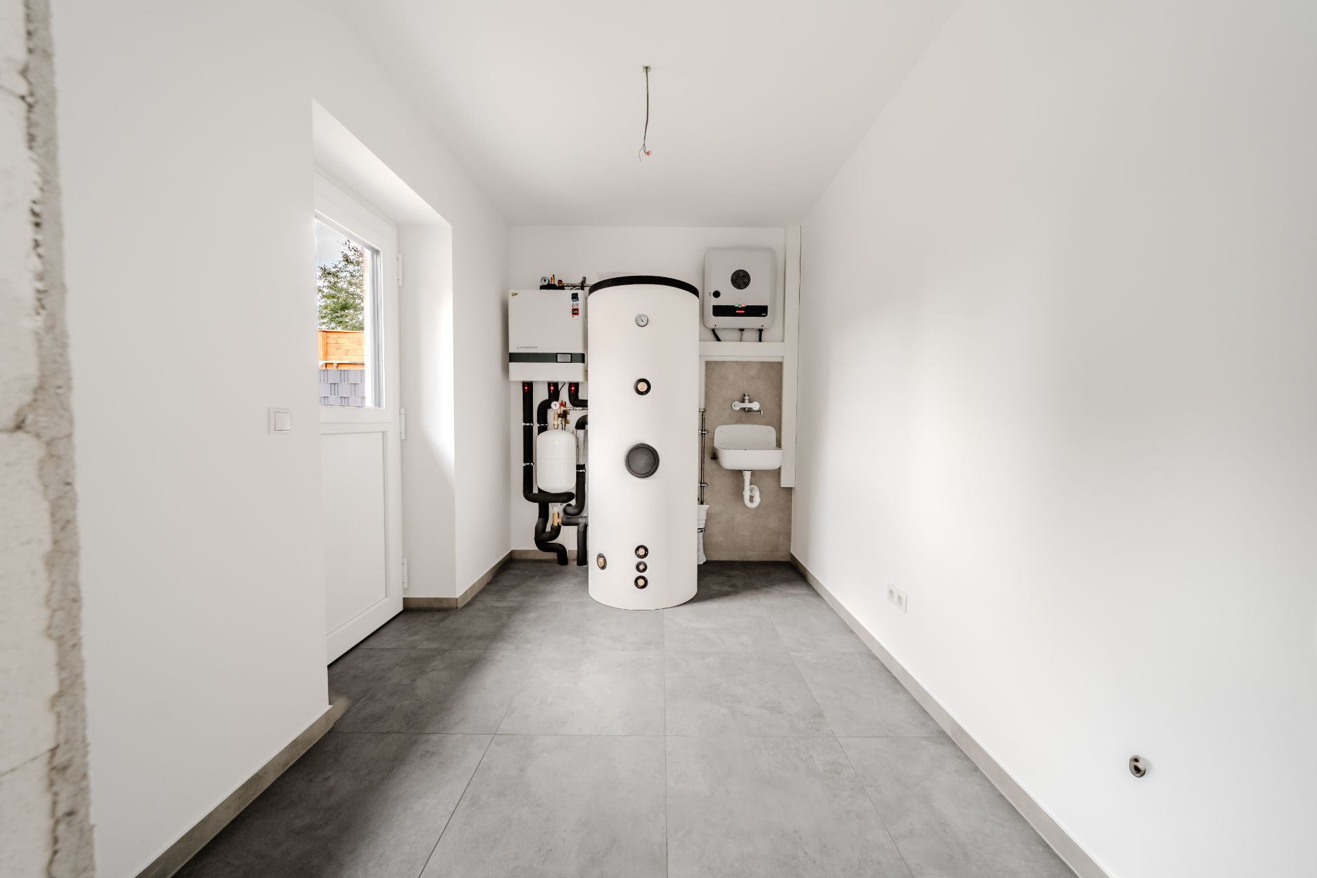 Hauswirtschaftsraum mit Boiler, Waschbecken und Heizanlage, weiße Wände, grauer Boden.