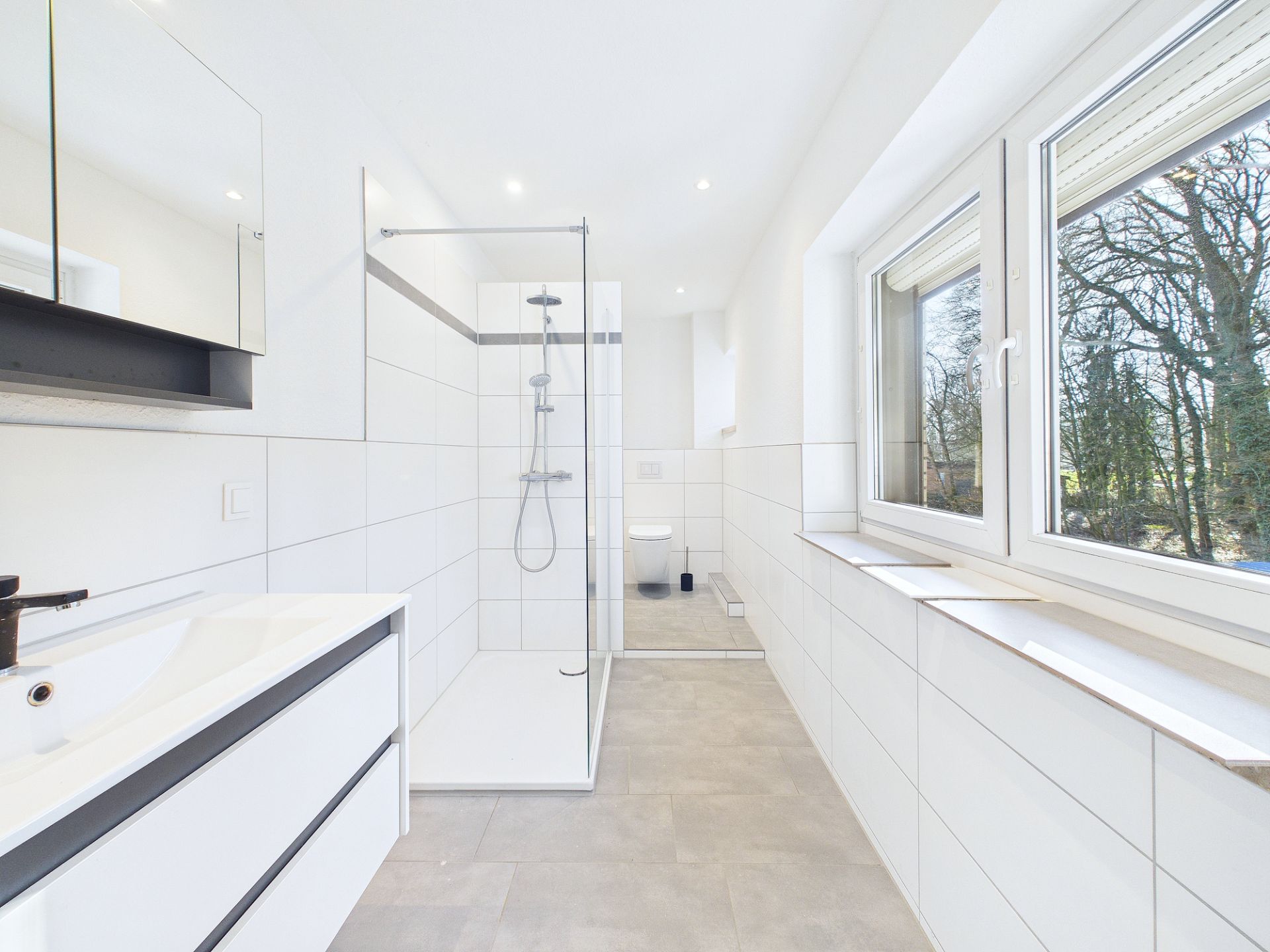 Modernes, weiß gefliestes Badezimmer mit großer Dusche, WC, Waschbecken und Fenster.