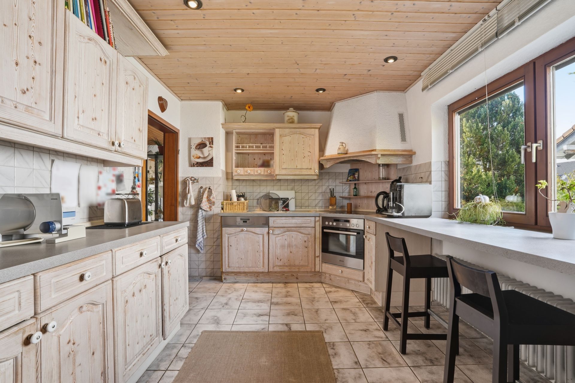 Helle Küche mit Holzschränken, großen Fenstern, Esstheke und zwei schwarzen Stühlen.