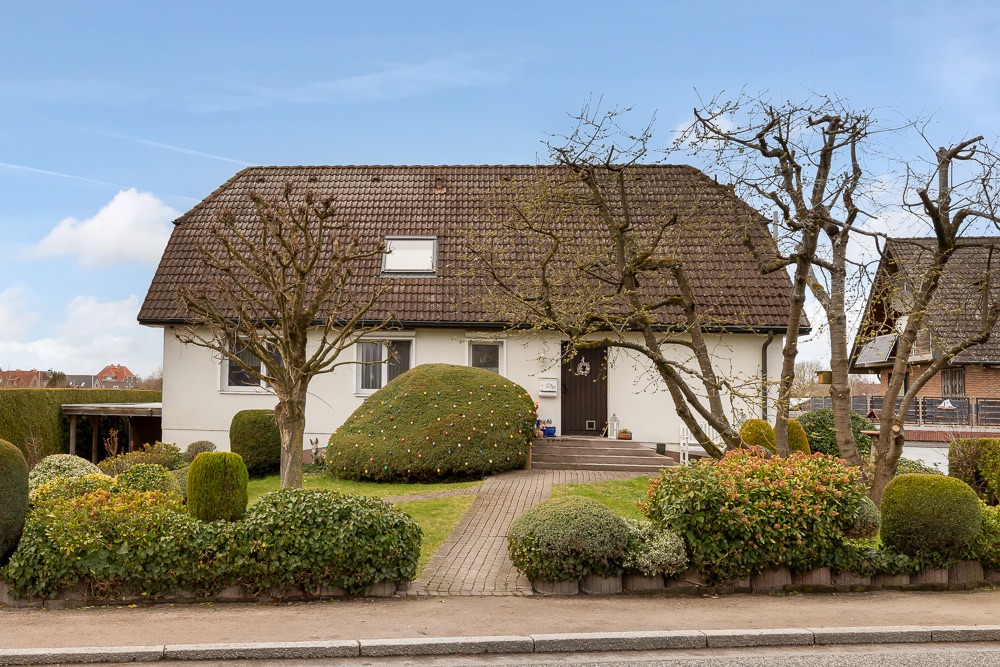 Einfamilienhaus mit braunem Dach, Vorgarten mit Büschen und Bäumen, gepflasterter Weg zum Eingang.