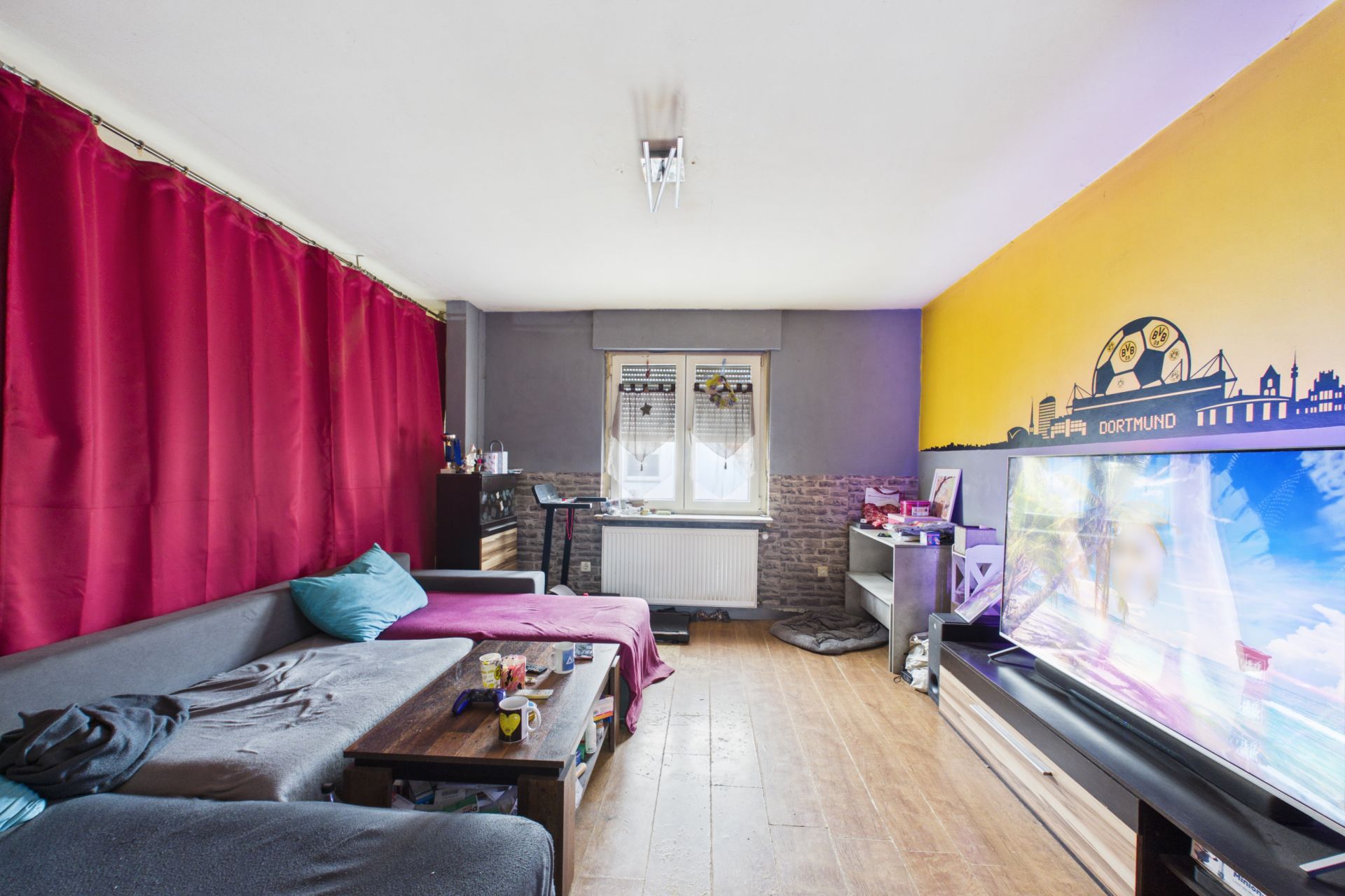 Wohnzimmer mit grauem Sofa, Couchtisch, rotem Vorhang und großem Fernseher an gelber Wand.