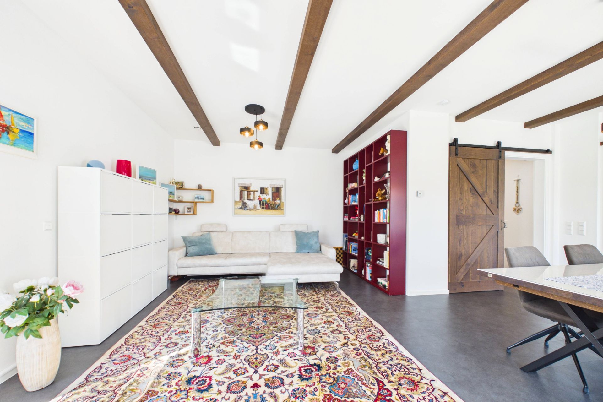 Helles Wohnzimmer mit weißem Sofa, Teppich, Bücherregal, Esstisch und Holzbalken an der Decke.